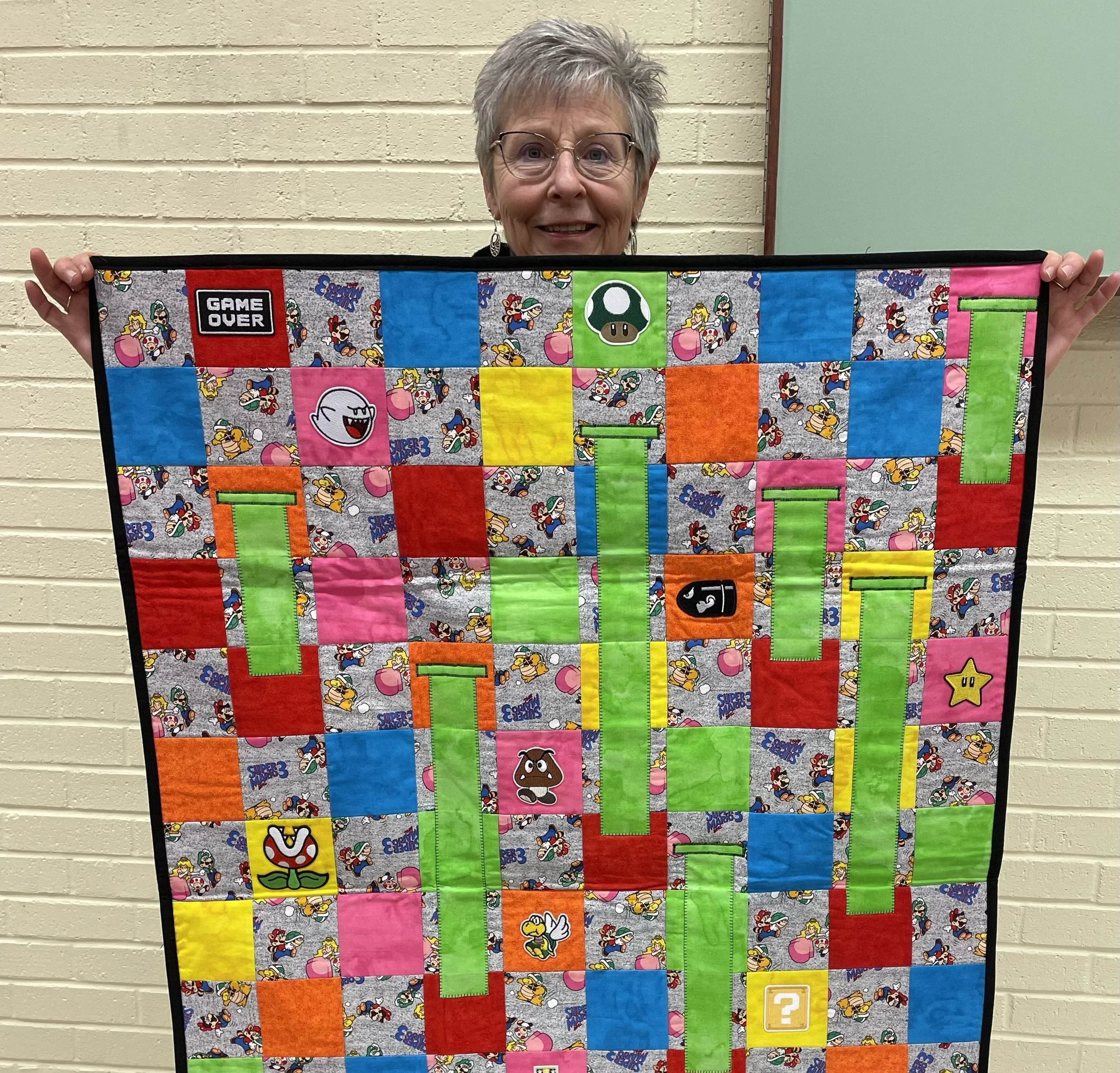 Person holding a quilt with Super Mario-themed design including characters like Boo, Goomba, and various game symbols on colorful patchwork squares.