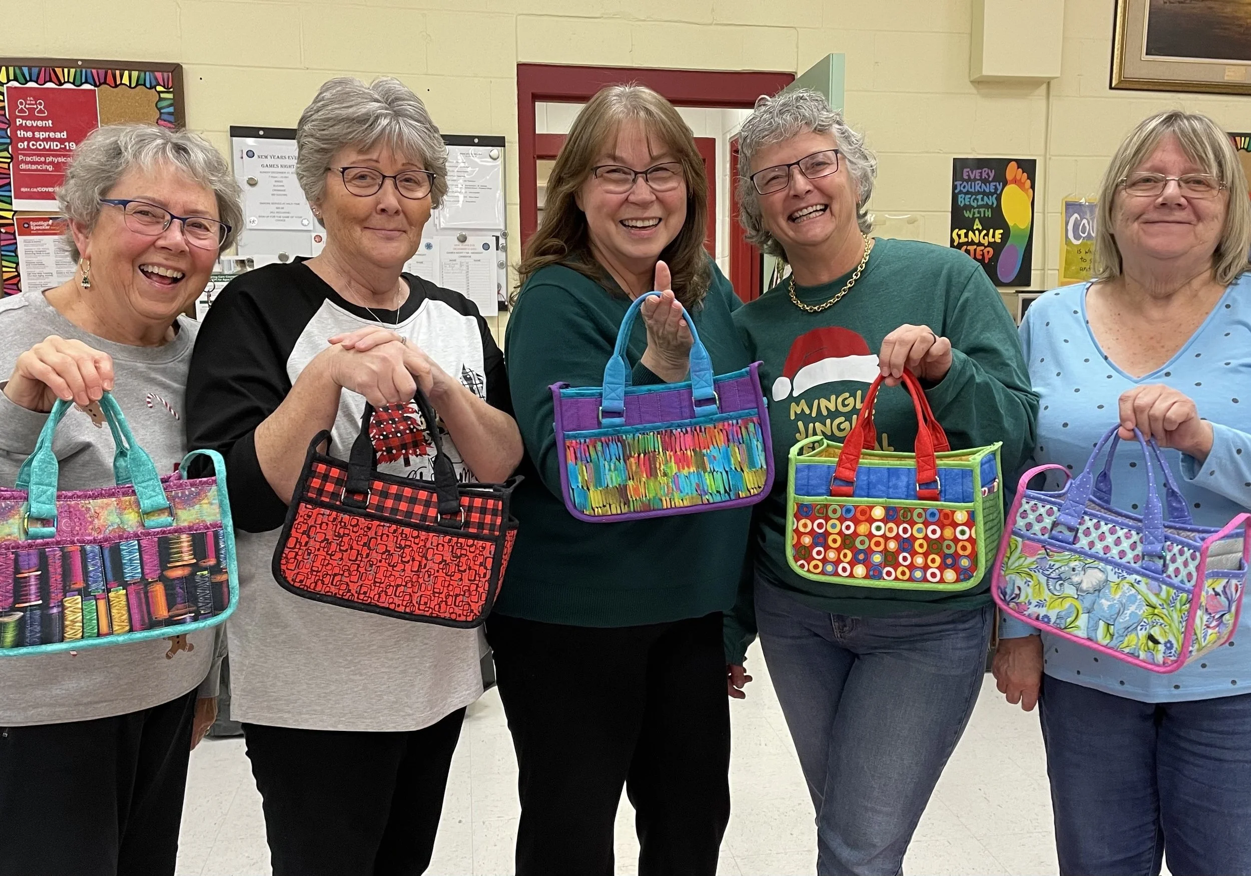 Five women smiling and holding colorful, handmade bags in an indoor setting.