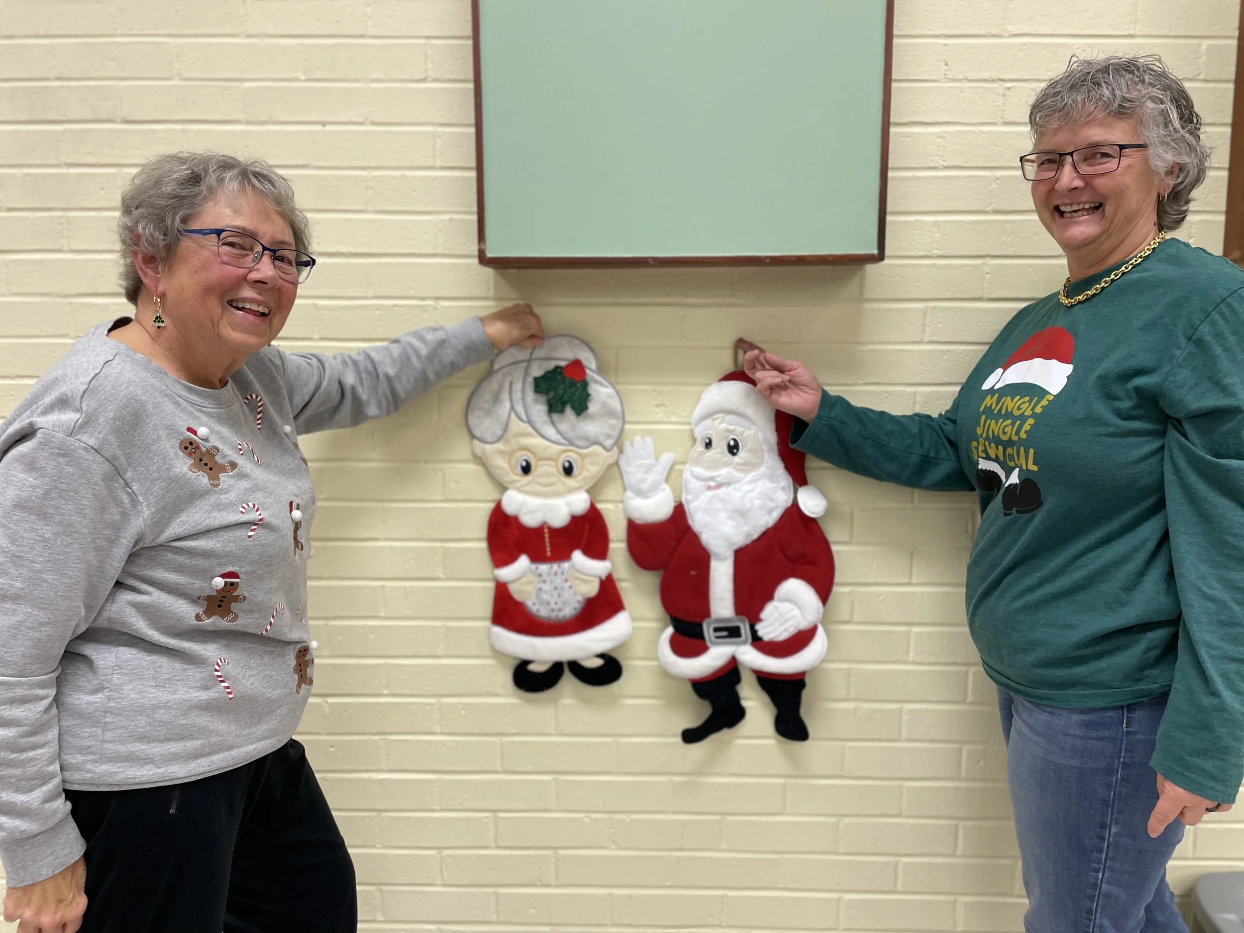 Two women smiling, standing in front of a wall with Christmas felt decorations of Santa Claus and Mrs. Claus. One woman wears a gray sweater with gingerbread and candy cane designs, and the other wears a green sweater with a Santa hat graphic.