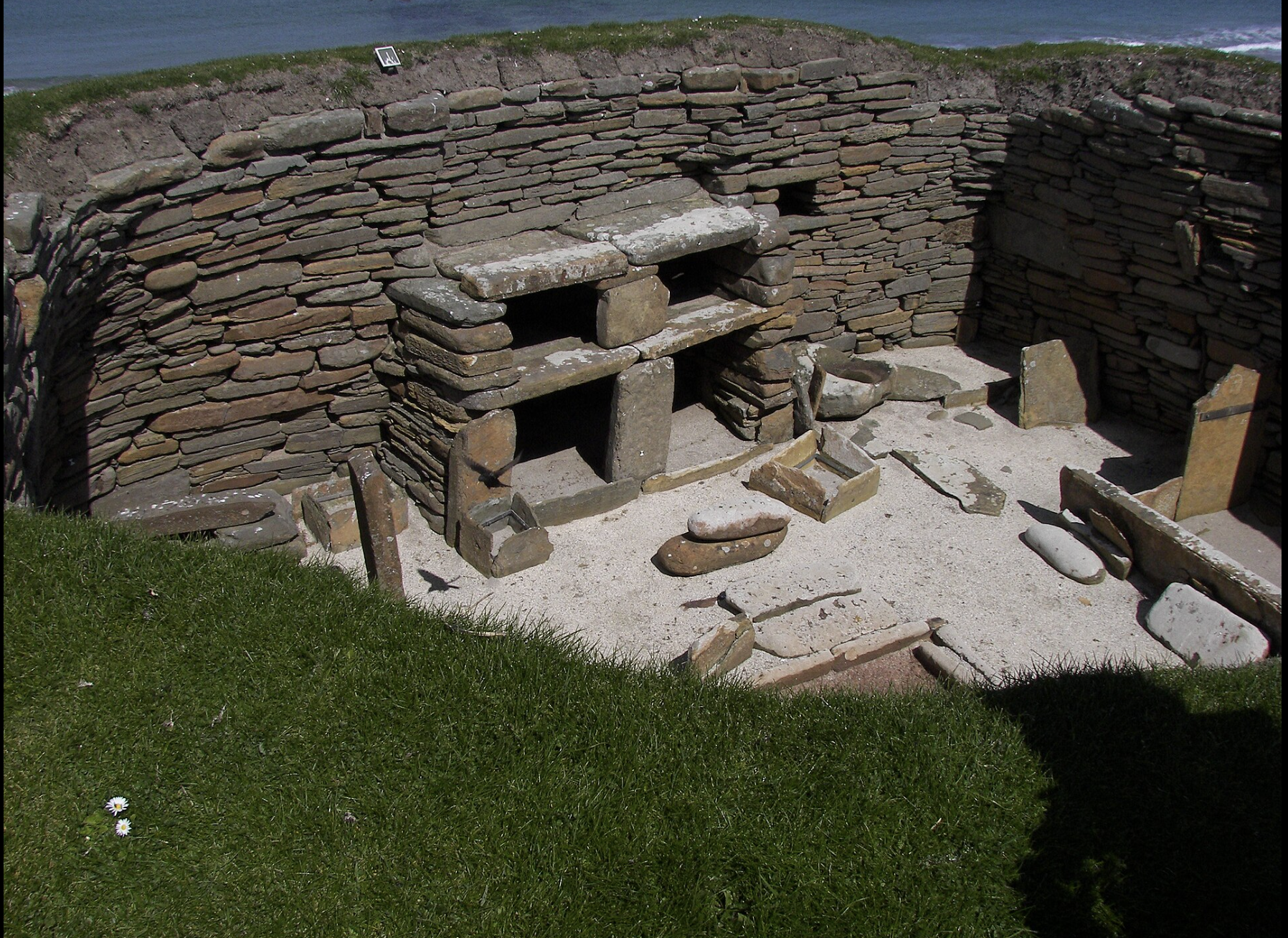 A dry stone structure, possibly a fireplace or firepit, built with stacked flat stones in a circular shape with open compartments, located outdoors on a sandy surface with grass in the foreground and a body of water in the background.