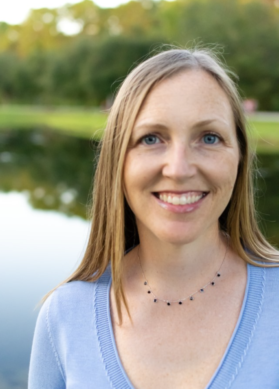 A smiling woman with blue eyes and light brown hair outdoors near a body of water with green trees in the background, wearing a light blue top and a black gemstone necklace.