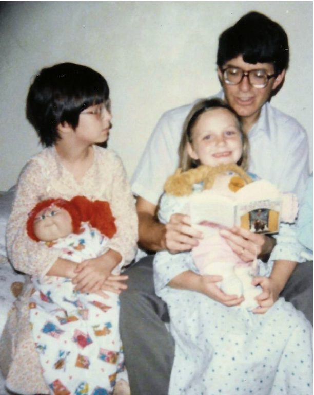 A man with dark hair and glasses sitting on a bed with two young girls, one of whom is holding a doll and a teddy bear, while the other girl is holding a book.
