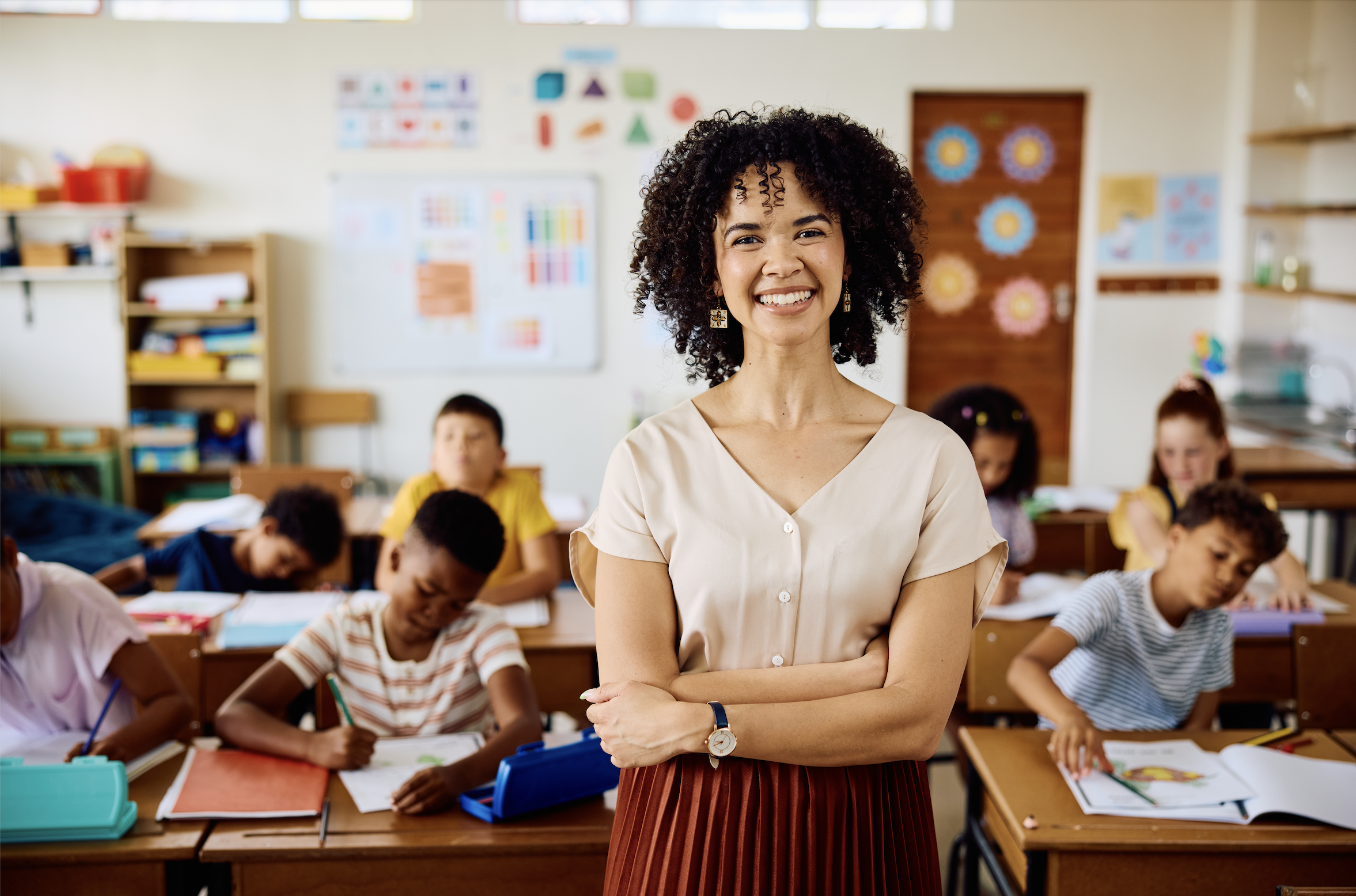 A smiling female teacher with curly hair and earrings standing in a classroom with students sitting at their desks doing work.