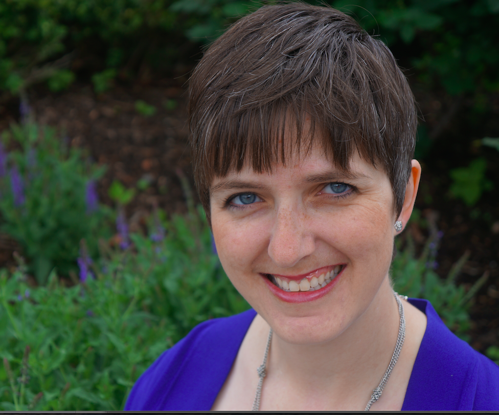 Close-up of a smiling woman with short brown hair and blue eyes outdoors.