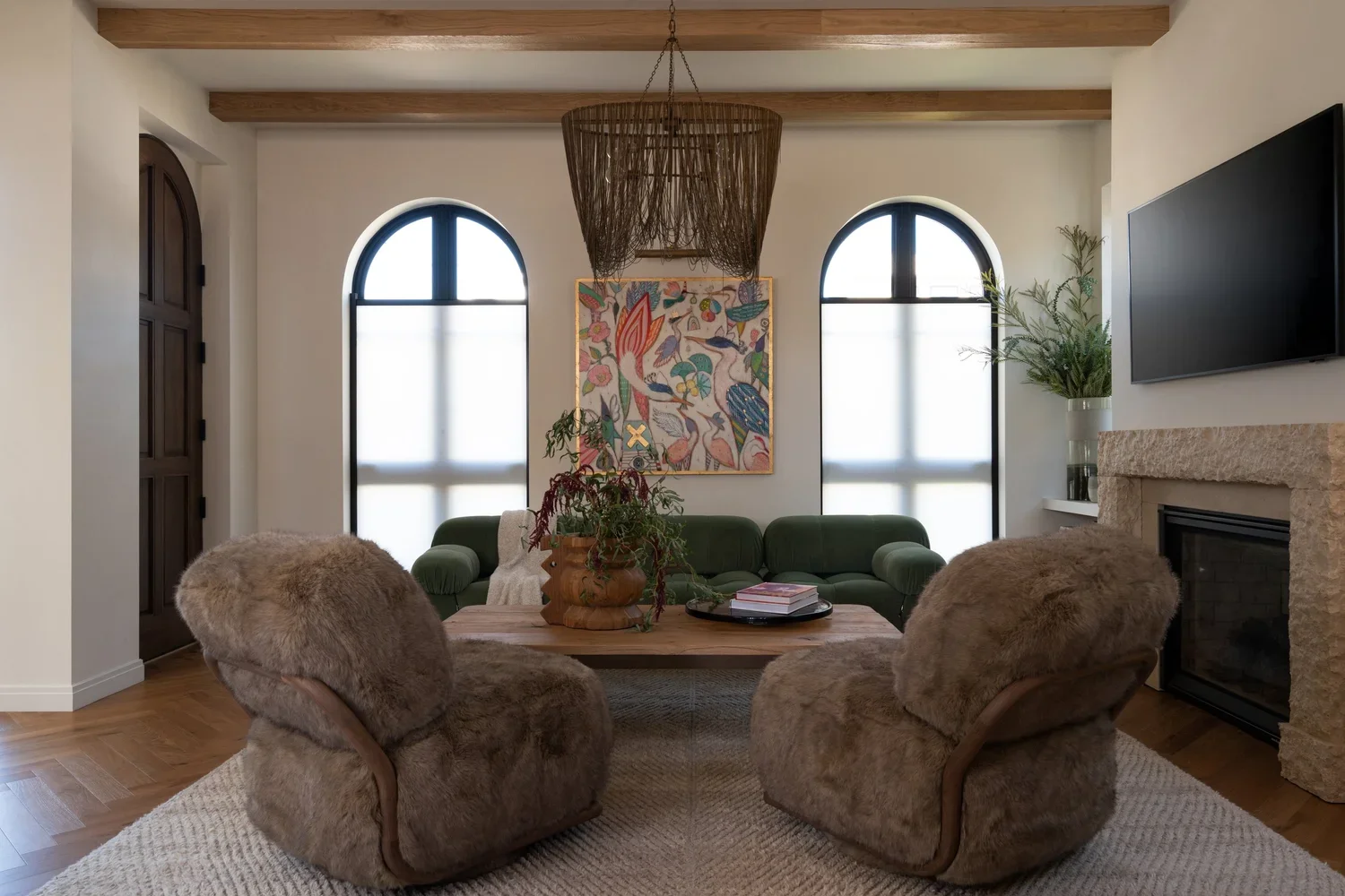 Living room with two fur-covered armchairs, a green sofa, and a wooden coffee table with plants and books. Bright windows with arched tops, a colorful artwork by Gretchen Weller Howard on the wall.