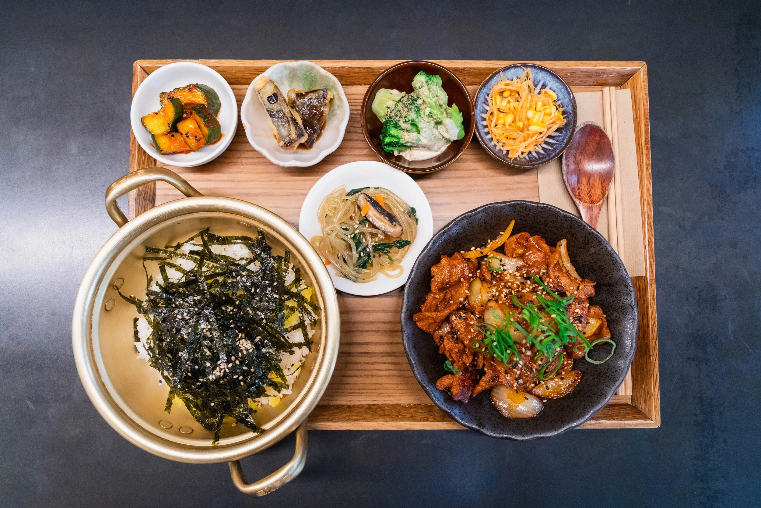 A traditional Korean meal served on a wooden tray with various side dishes, a main dish of spicy stir-fried meat and vegetables, and a bowl of rice topped with seaweed strips.