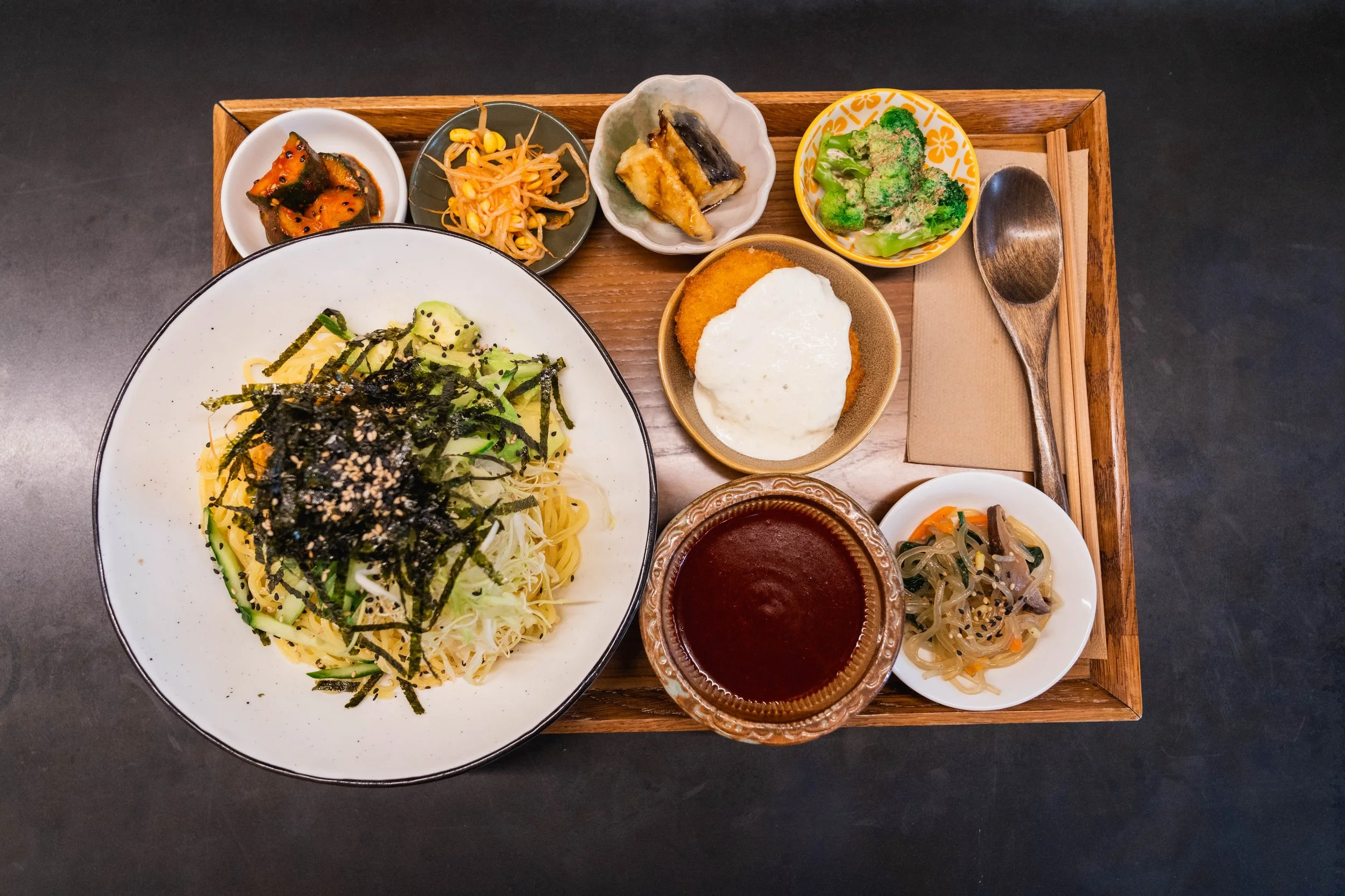 Top-down view of a Japanese meal with various dishes and condiments on a wooden tray. Includes a large bowl of spaghetti topped with seaweed strips, several small side dishes with vegetables and fried items, a glass of dark beverage, and a spoon with chopsticks on the right side.