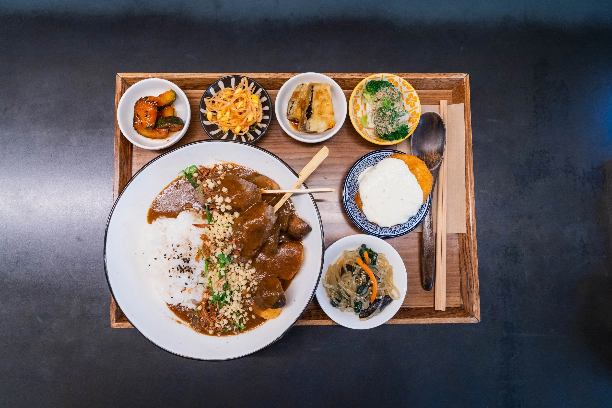 A Korean meal served on a wooden tray, featuring a large bowl of curry rice with chicken and toppings, surrounded by small side dishes including pickles, salad, grilled fish, broccoli, and mixed vegetables, with a spoon and chopsticks.