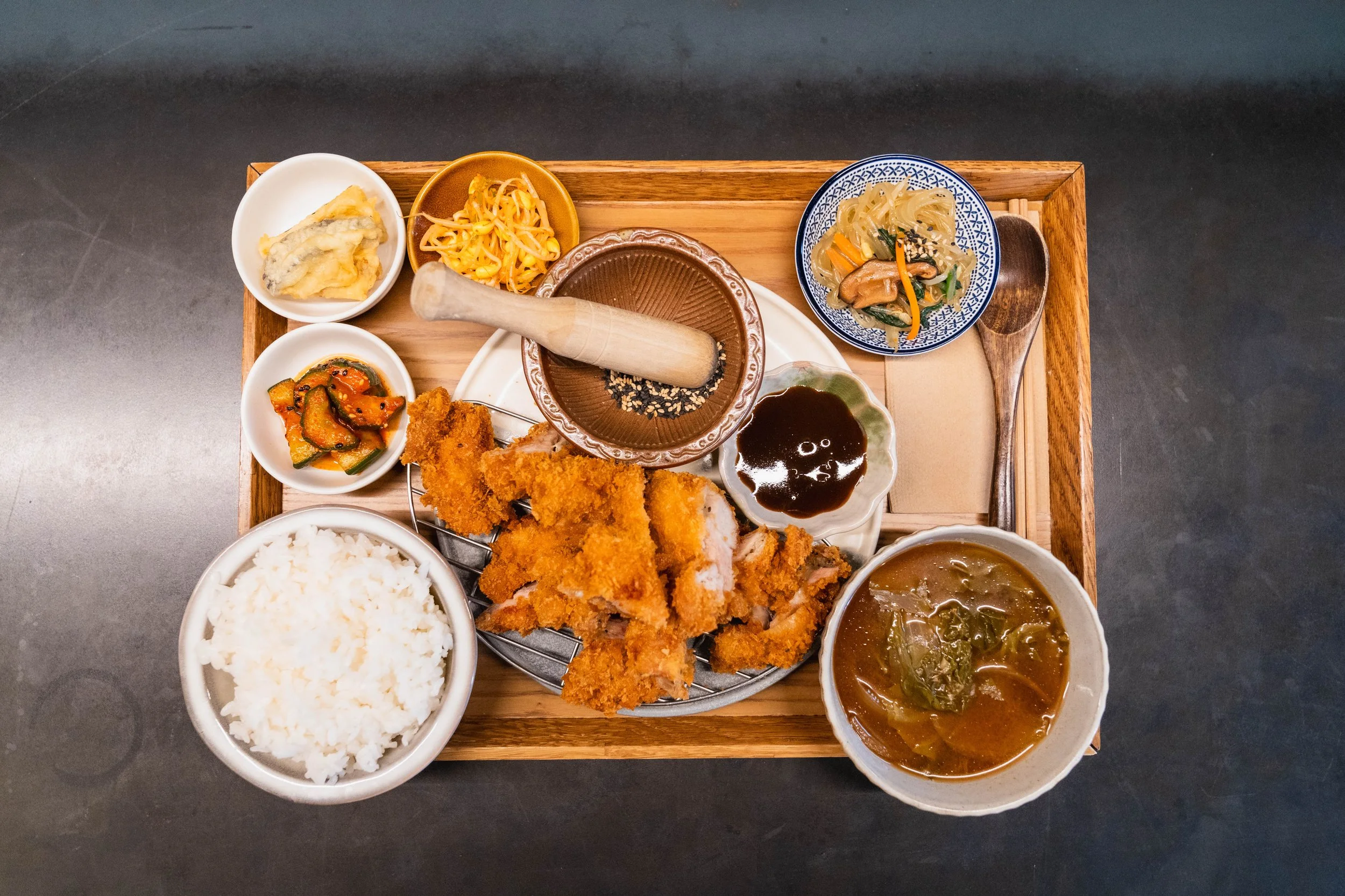 Top-down view of a Korean meal served on a wooden tray, including rice, fried chicken, various side dishes, dipping sauces, and soup.