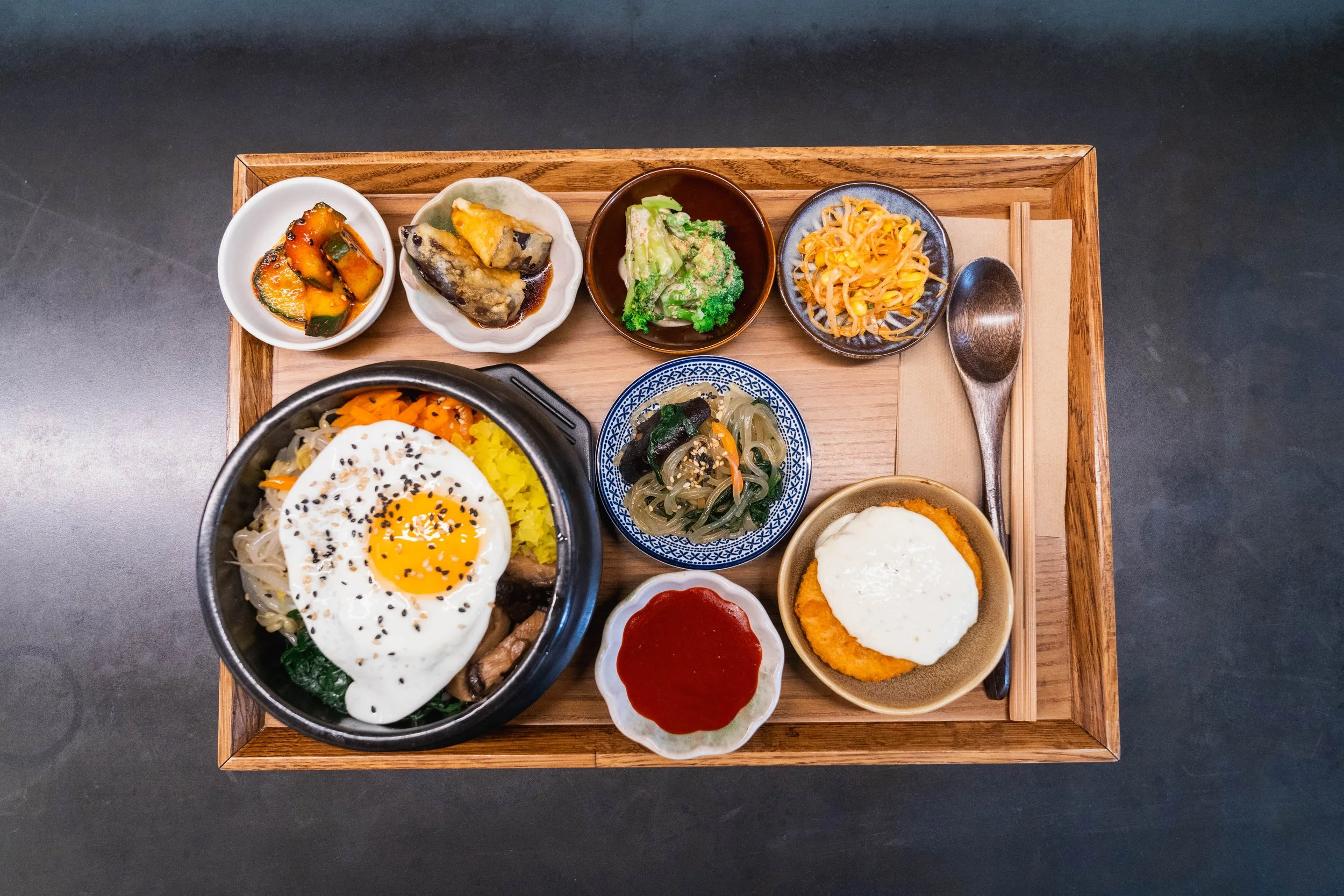 A tray of Korean cuisine with bibimbap topped with a fried egg, accompanied by various side dishes including kimchi, cucumber, broccoli, bean sprouts, seaweed salad, and a serving of fried chicken.