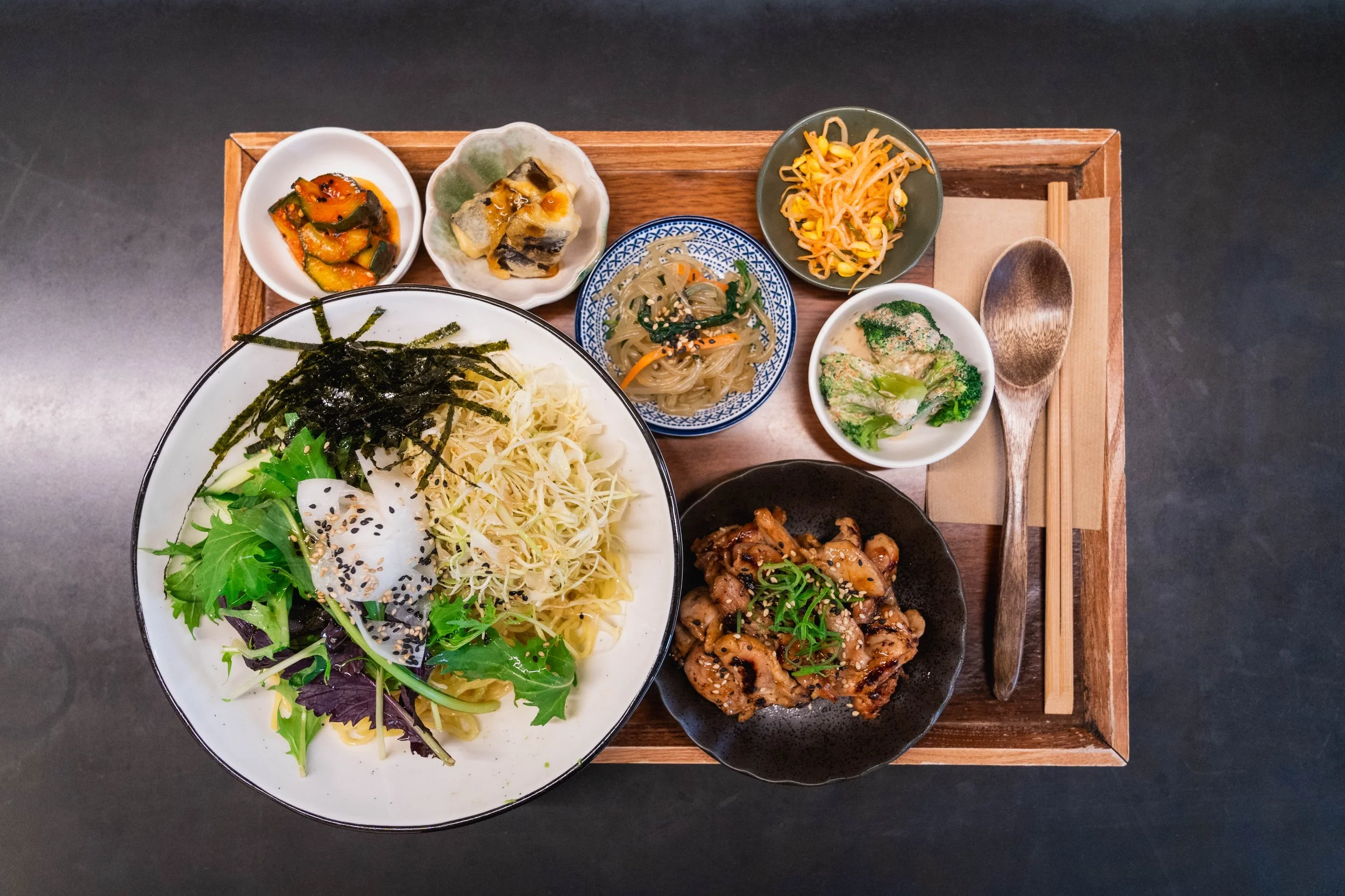 A traditional korean meal featuring a large bowl of salad with mixed greens, shredded chicken, seaweed, and sesame seeds, accompanied by smaller dishes of pickled vegetables, marinated seaweed, bean sprouts, broccoli, grilled chicken