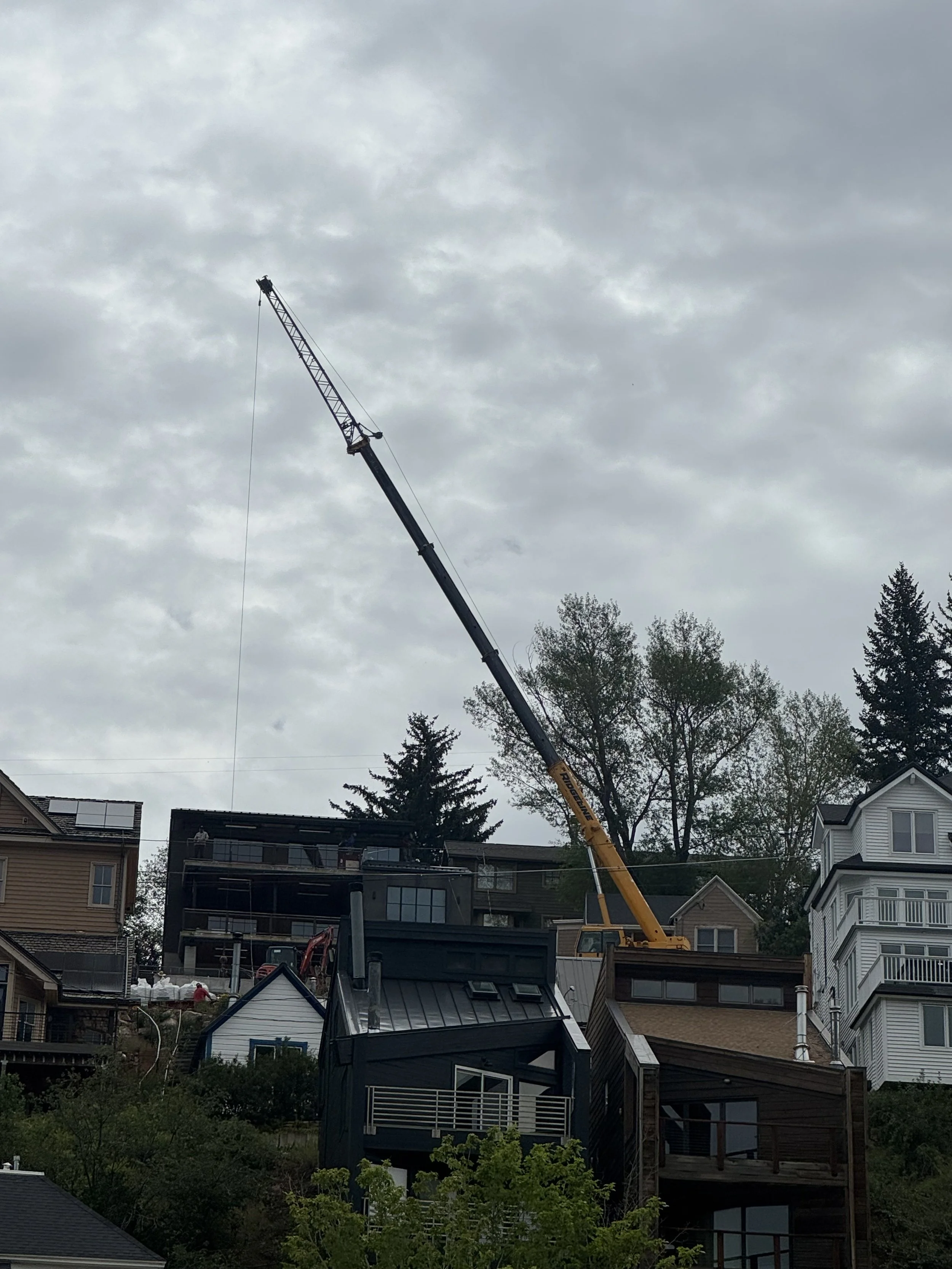 A construction crane working in a neighborhood with multiple residential buildings and trees under a cloudy sky.