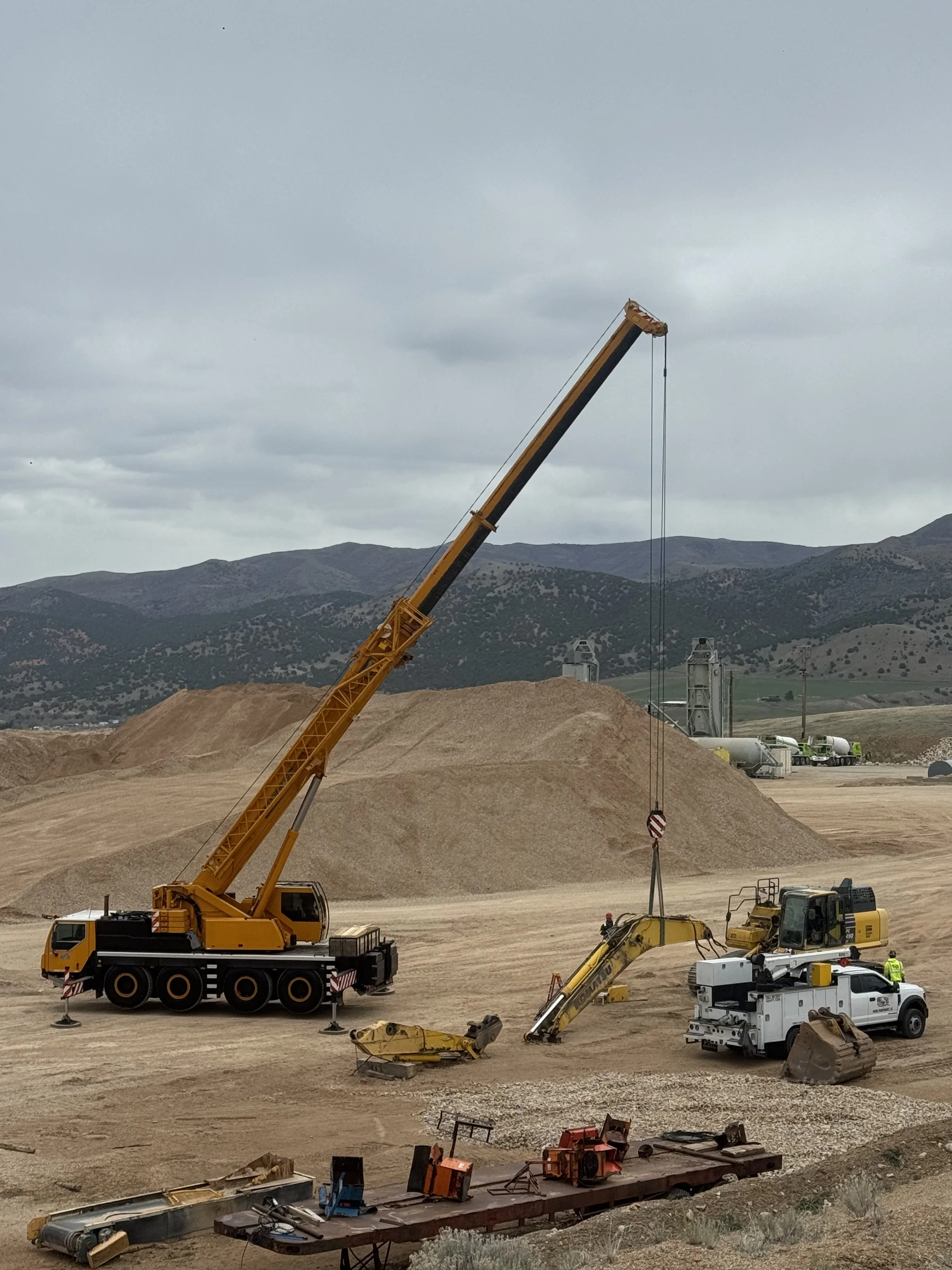 Construction site with a large yellow crane lifting equipment or materials, surrounded by other heavy machinery and trucks, in a dirt area with mountains and overcast sky in the background.