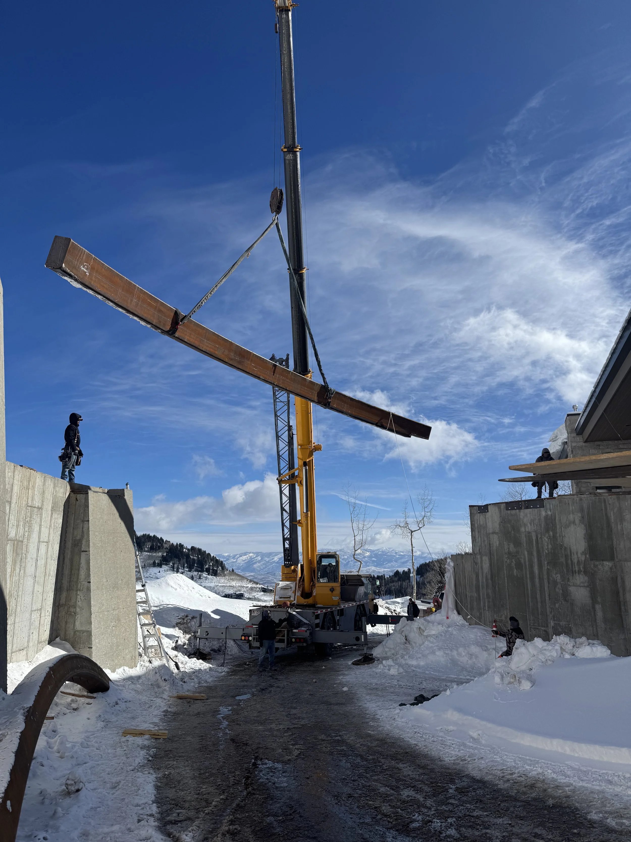 Construction workers operate a crane installing a large wooden beam in a snowy mountain setting with concrete walls on either side.