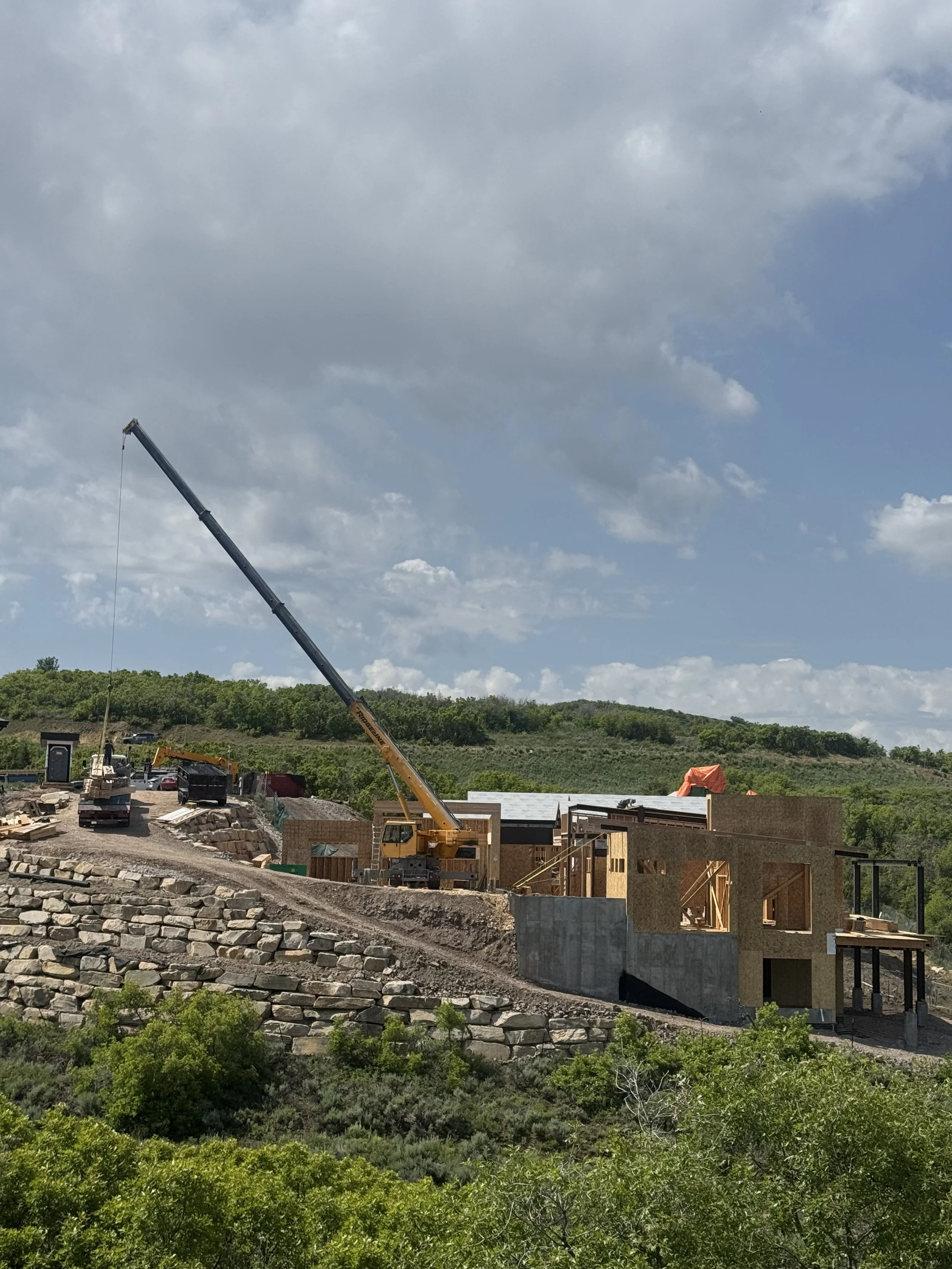 Construction site on a hillside with a crane, partially built structures, and surrounding greenery under a blue sky with clouds.
