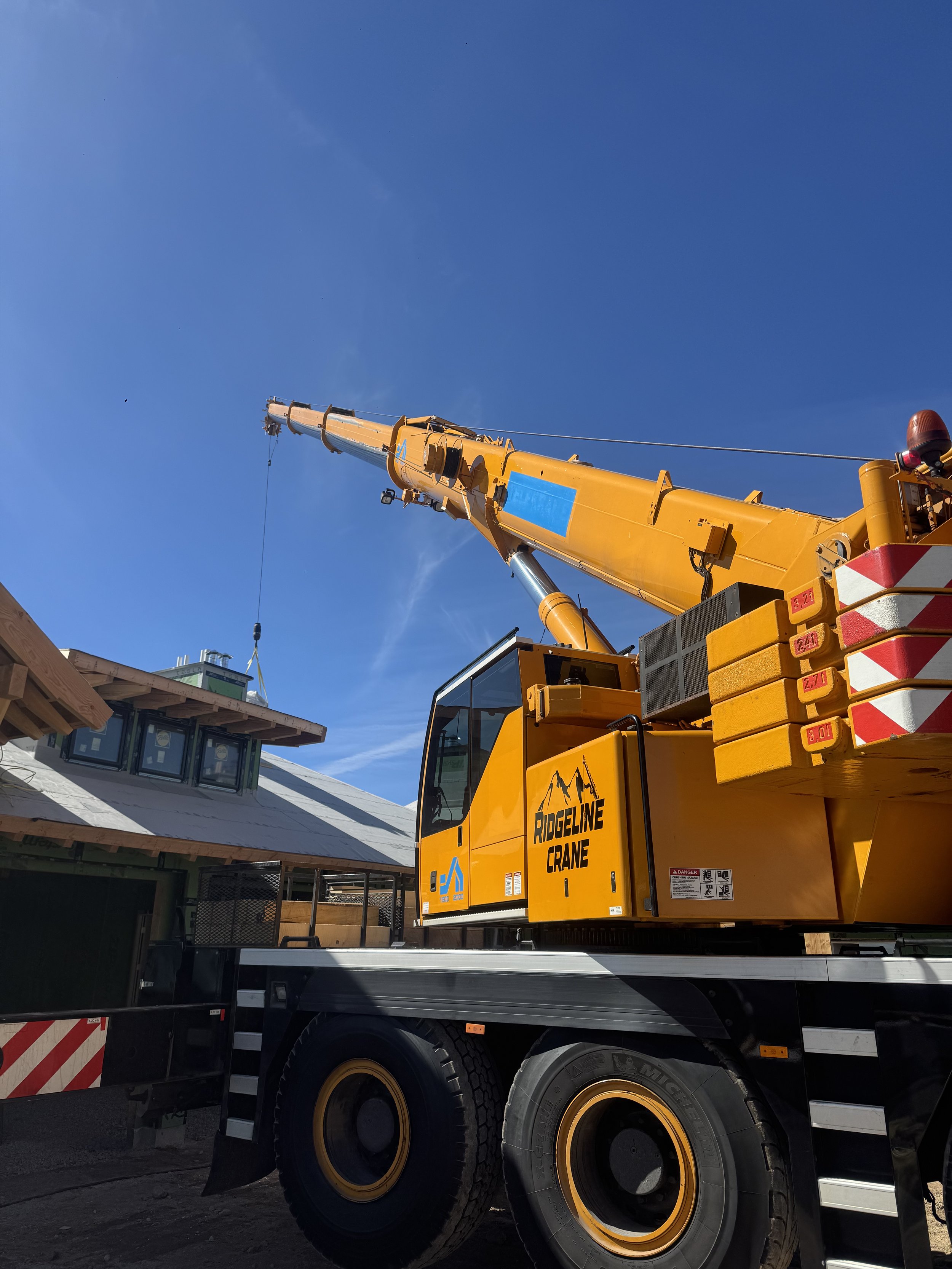 A large yellow crane truck, labeled 'RIDGELINE CRANE,' is positioned at a construction site under a clear blue sky. The crane's arm is extended upward, and the site includes a partially constructed building with a metal roof and windows.