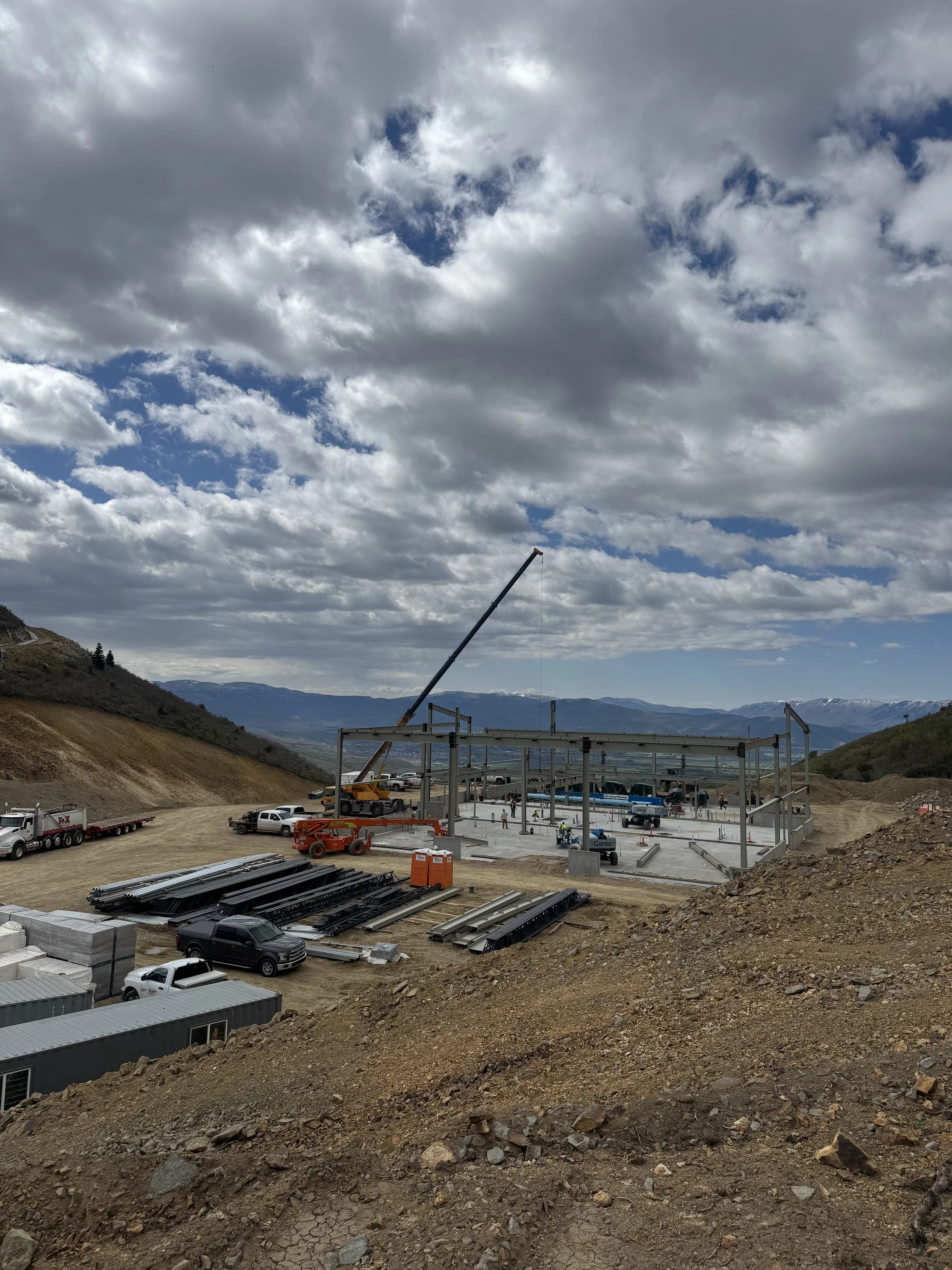 Construction site with partially built structure, construction equipment, and trucks, set against a mountainous landscape with cloudy sky.