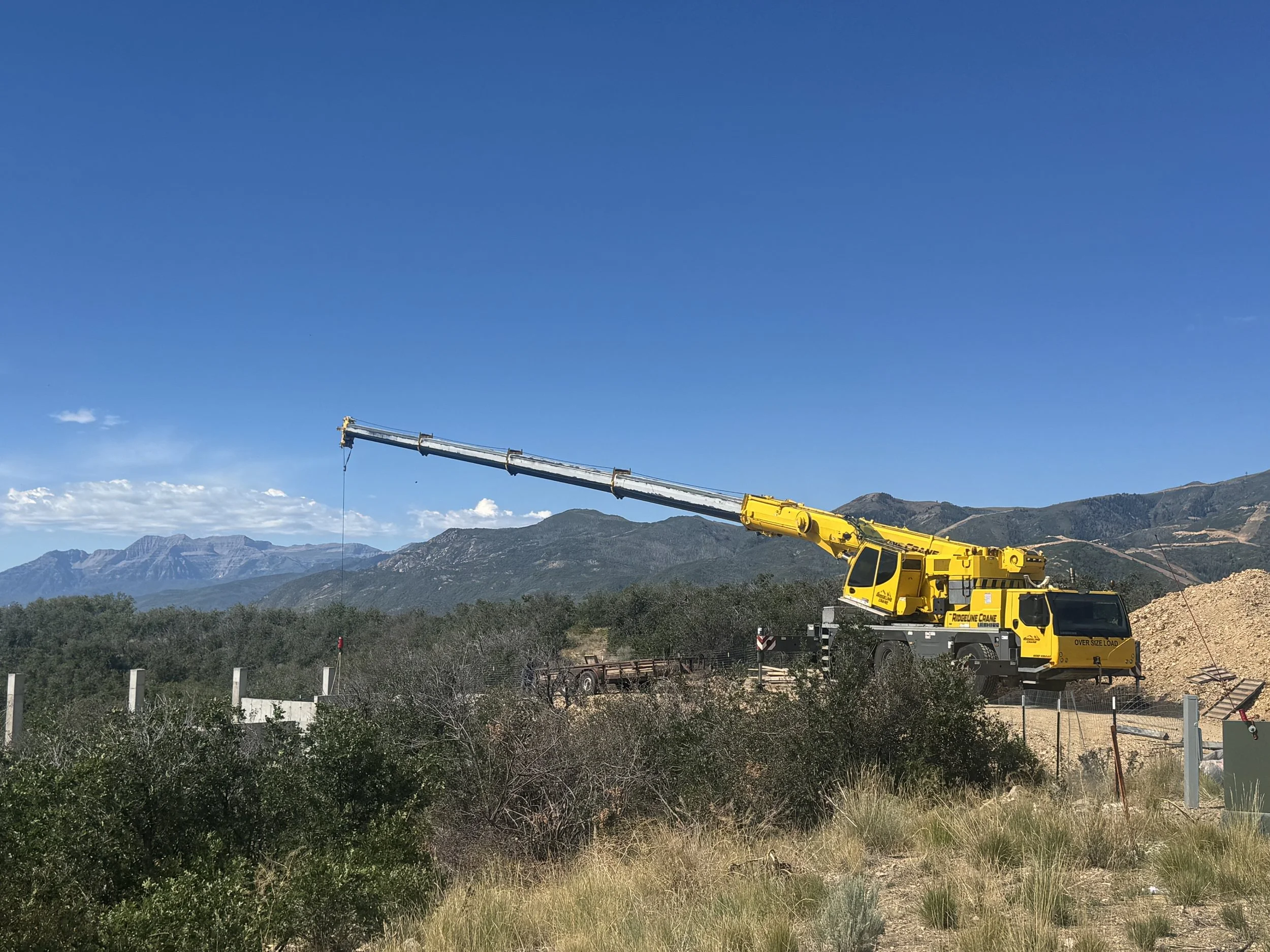 A yellow crane truck is operating on a construction site in a mountainous area, with mountains and a blue sky in the background.