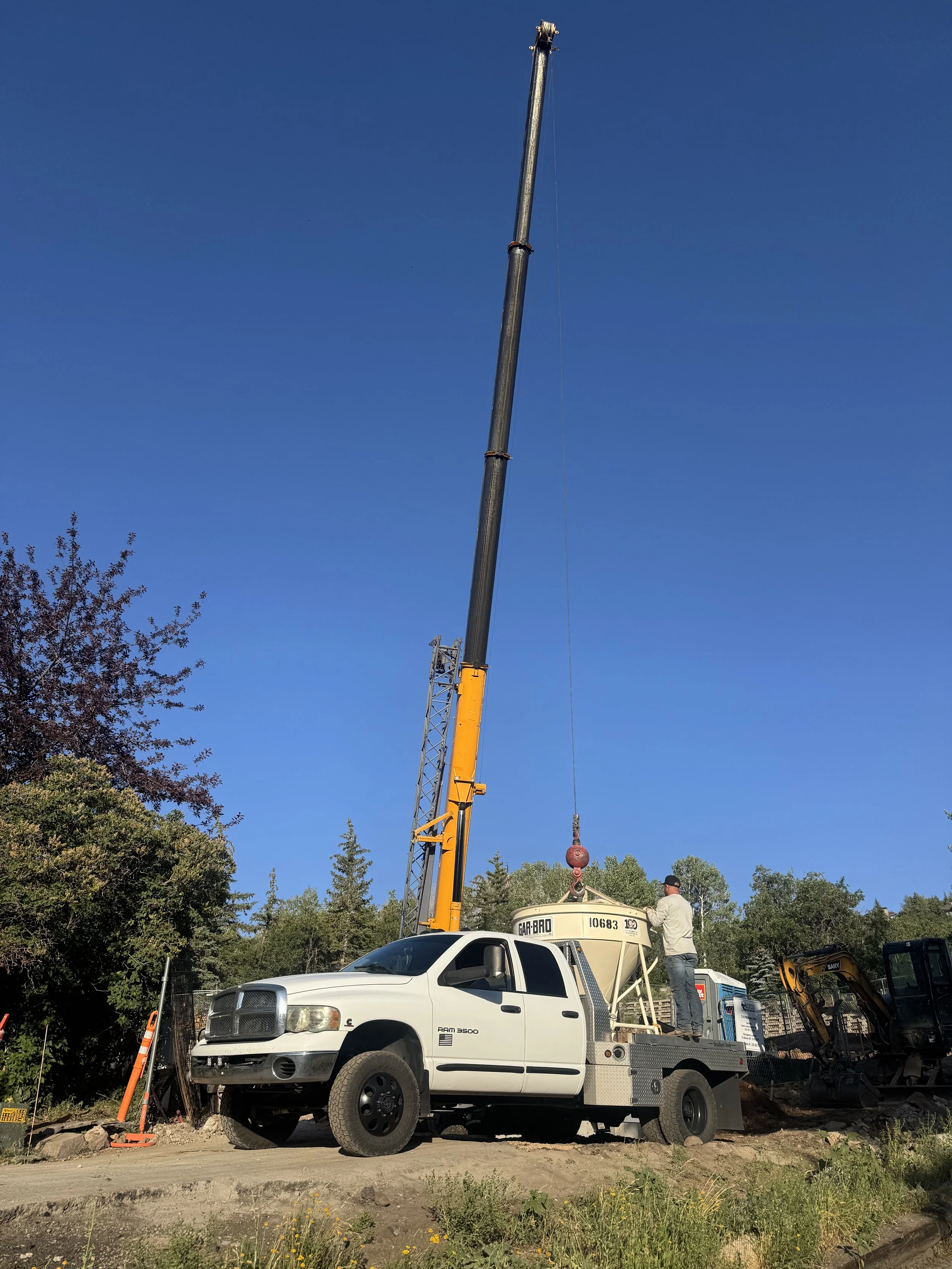 Construction crane lifting a large cement bucket with a worker standing nearby, a pickup truck parked in the foreground, surrounded by trees and a clear blue sky.