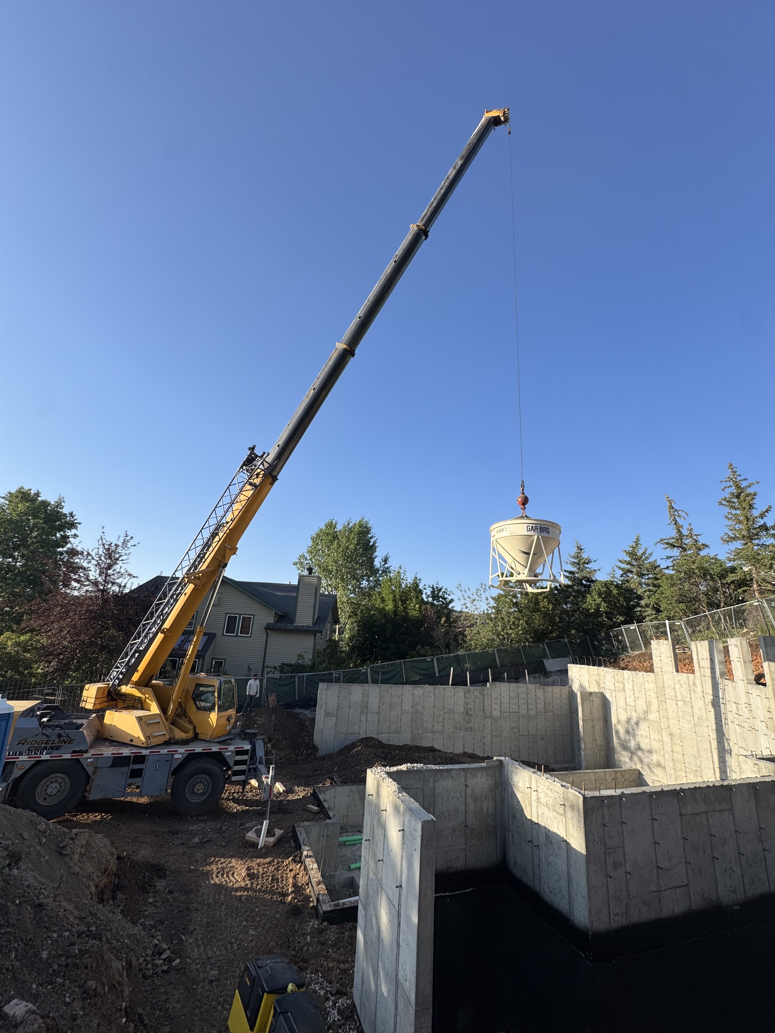 Construction site with a crane lifting a large concrete water tank into an underground foundation. The site is surrounded by a concrete wall and has trees and houses in the background.