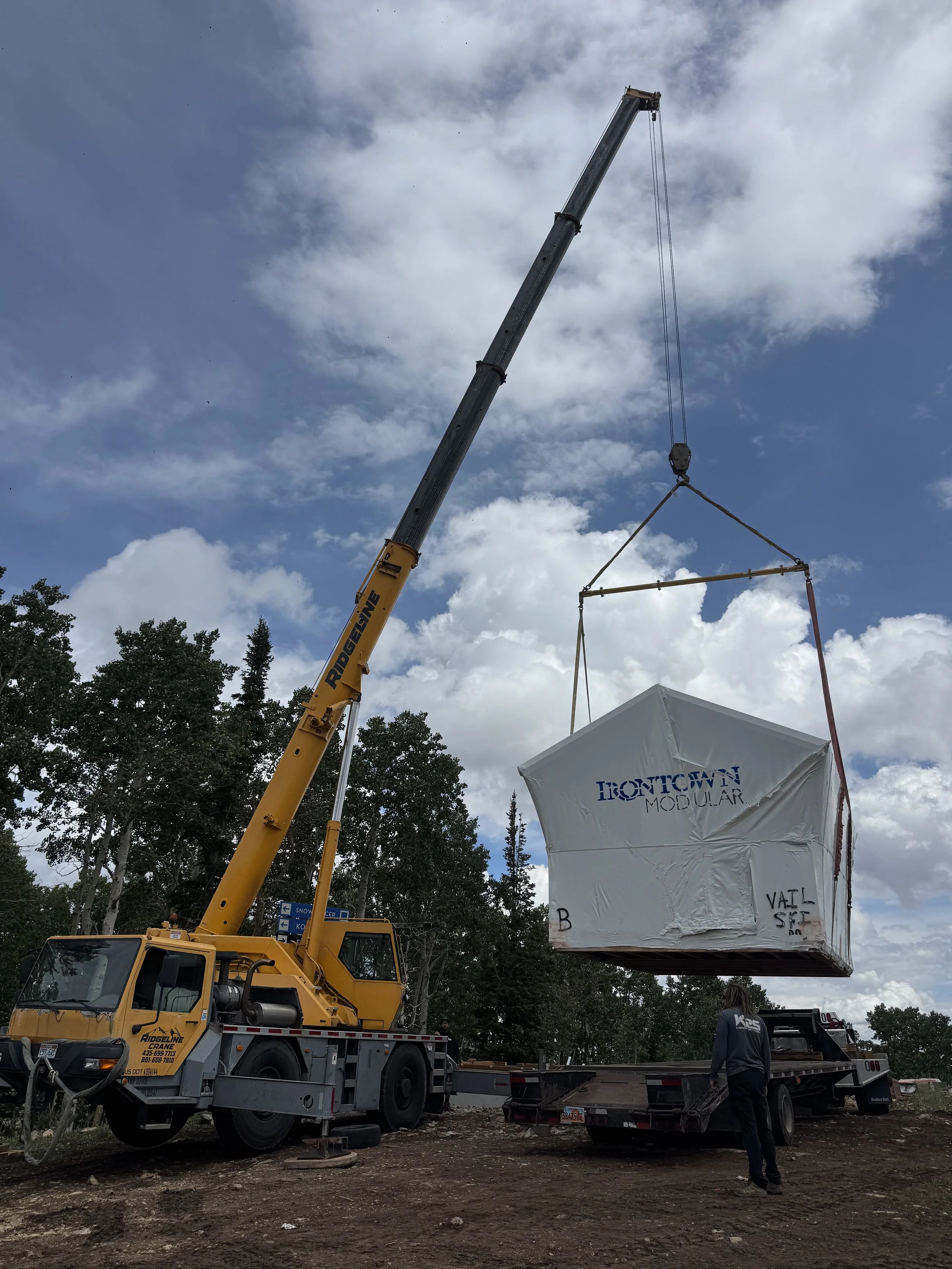 A large yellow crane lifting a wrapped structure labeled 'Frontier Modular' at a construction site with trees and a cloudy sky in the background.