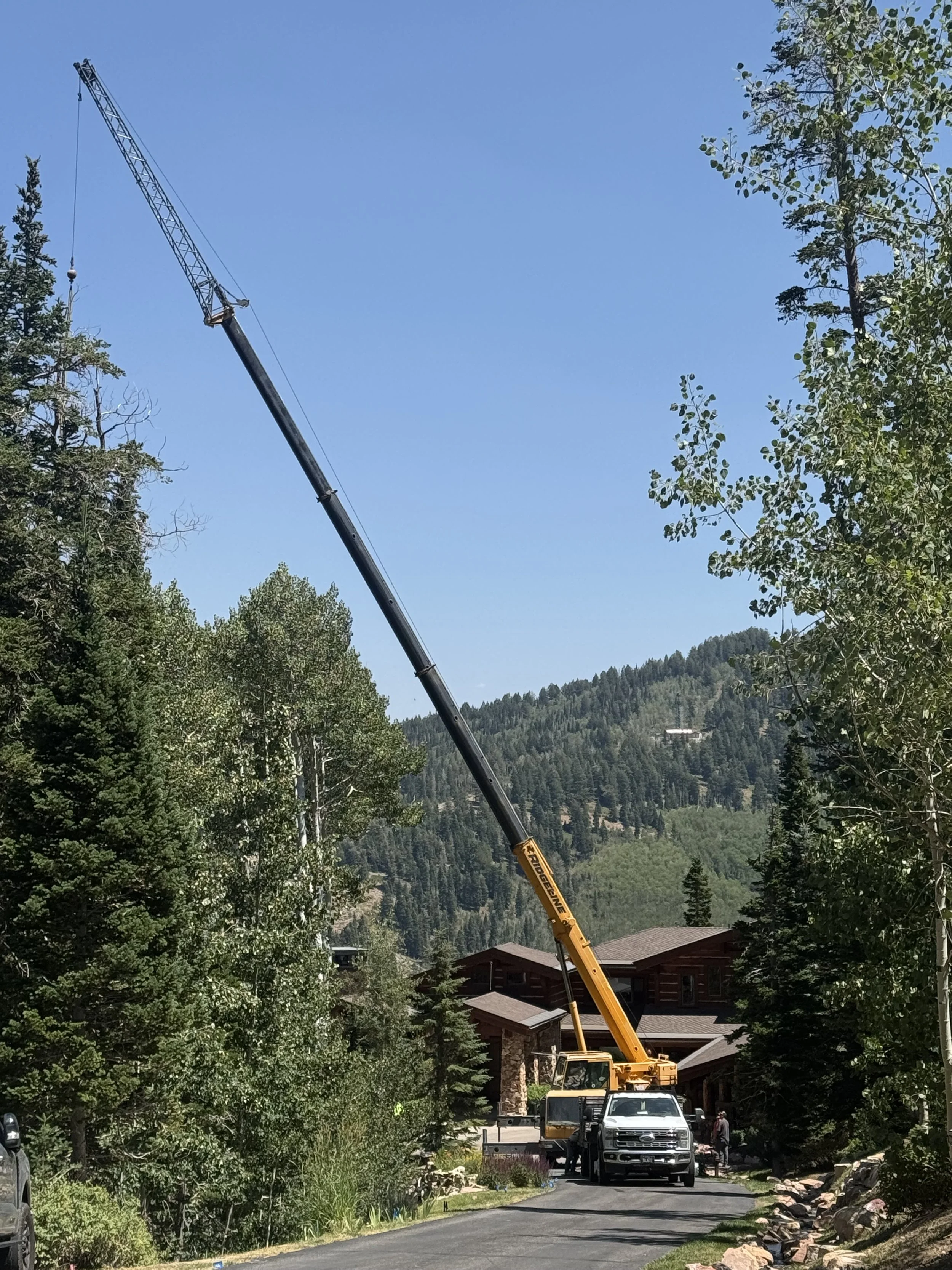 A yellow crane is lifting a large object between trees and houses with mountains in the background on a sunny day.