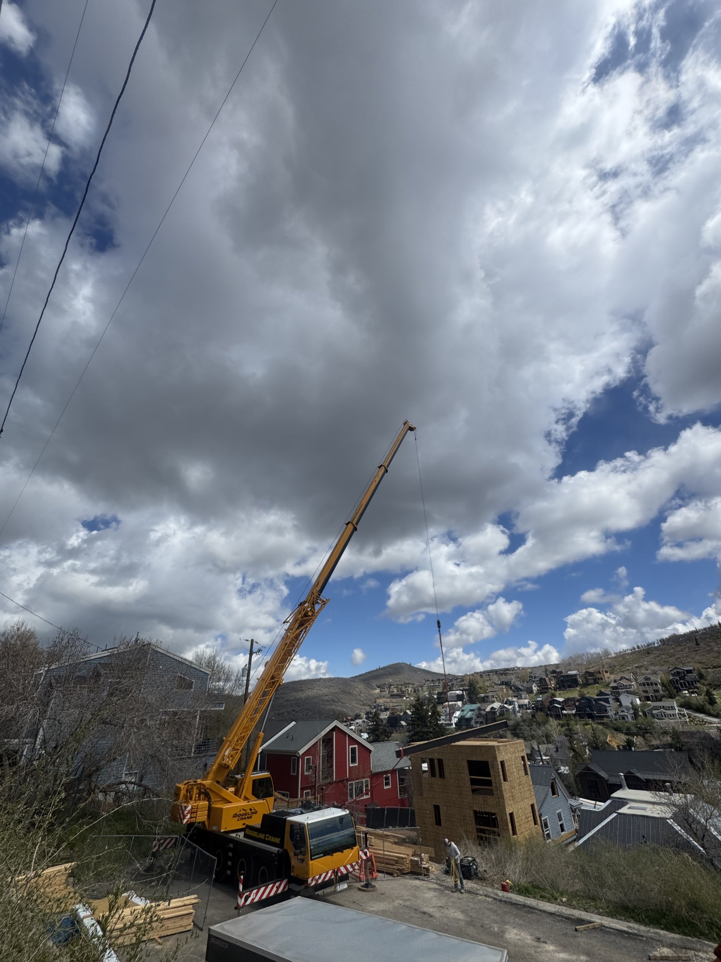 A large yellow crane is lifting a construction material over a neighborhood with houses and hillside, under cloudy skies.