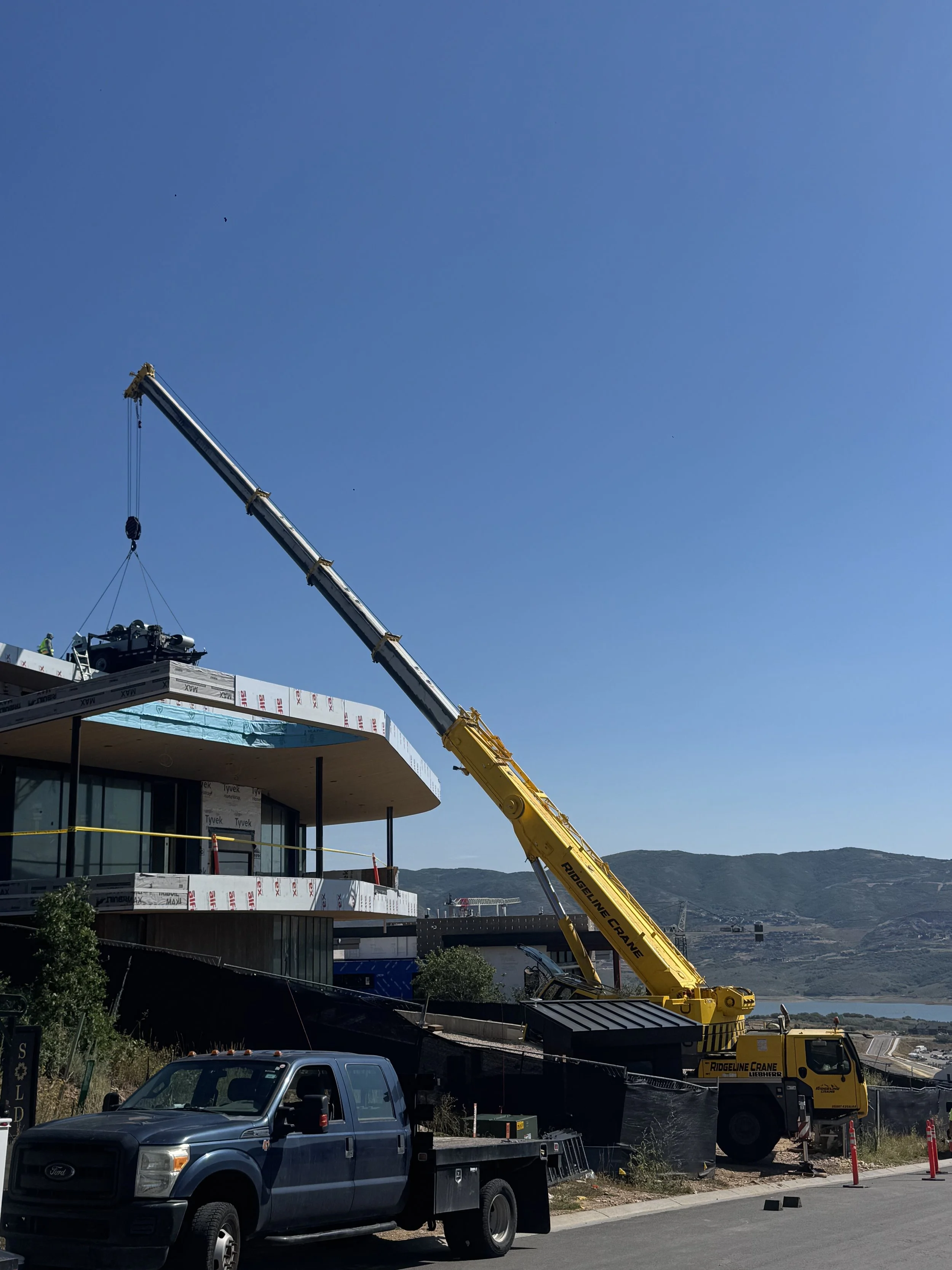 Construction site with a large yellow crane and a building under construction, with mountains in the background and a clear blue sky.