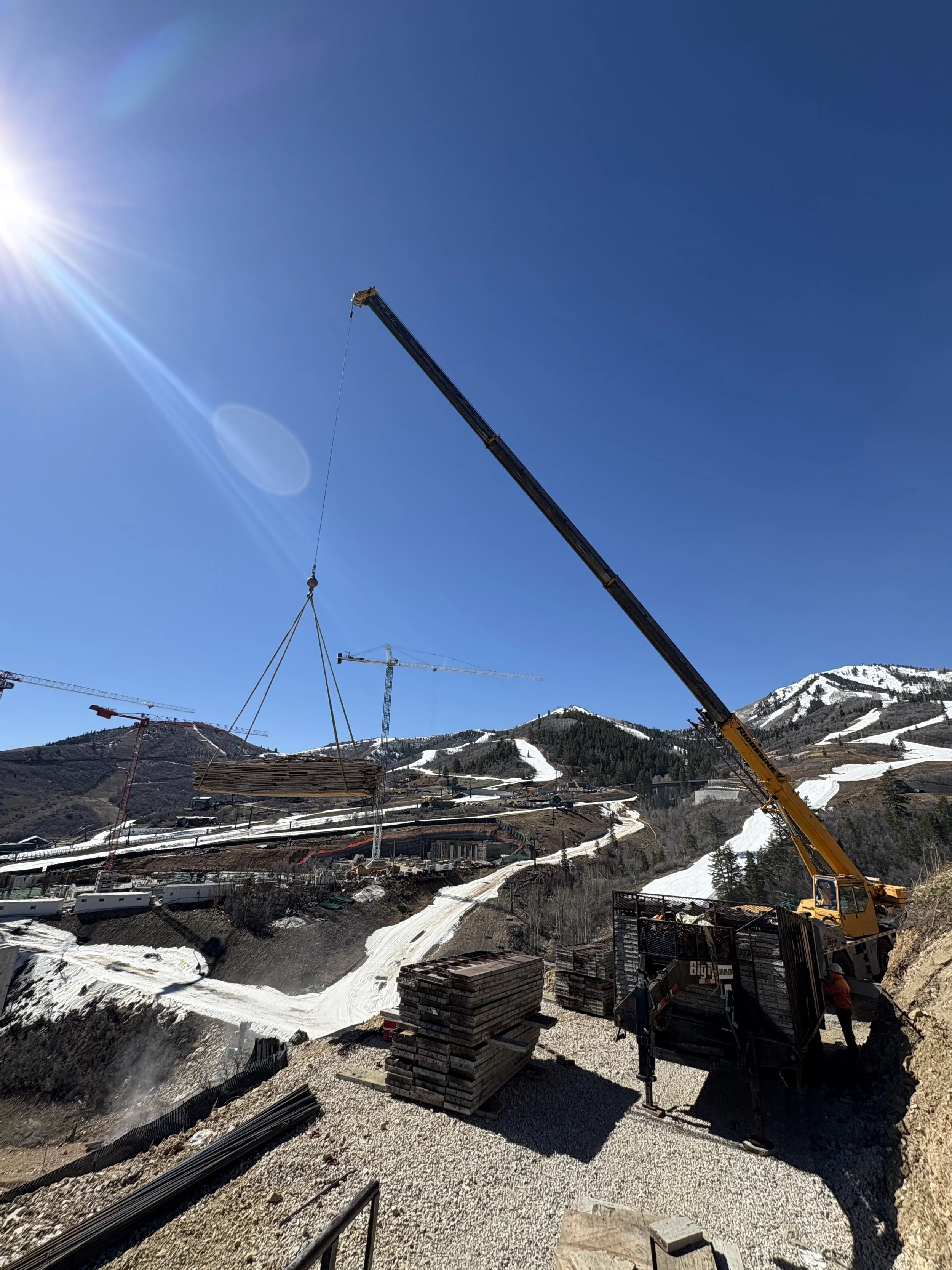 Construction site on a mountain with cranes and building materials, snow patches on the ground, and a clear blue sky with the sun shining.