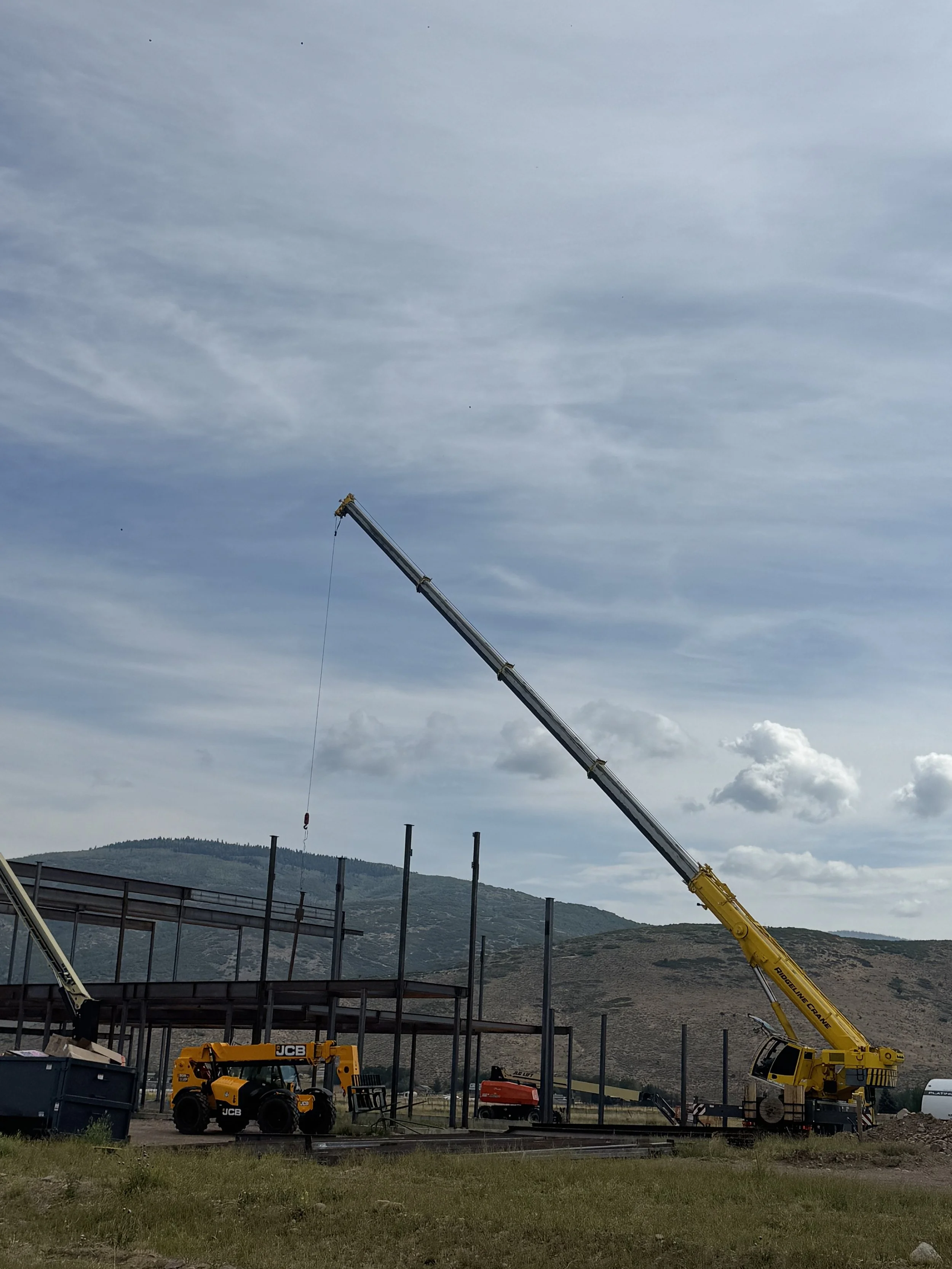 Construction site with a large yellow crane extending diagonally across the image and a smaller JCB machine in the foreground, with structural framework being built and mountains in the background under a cloudy sky.