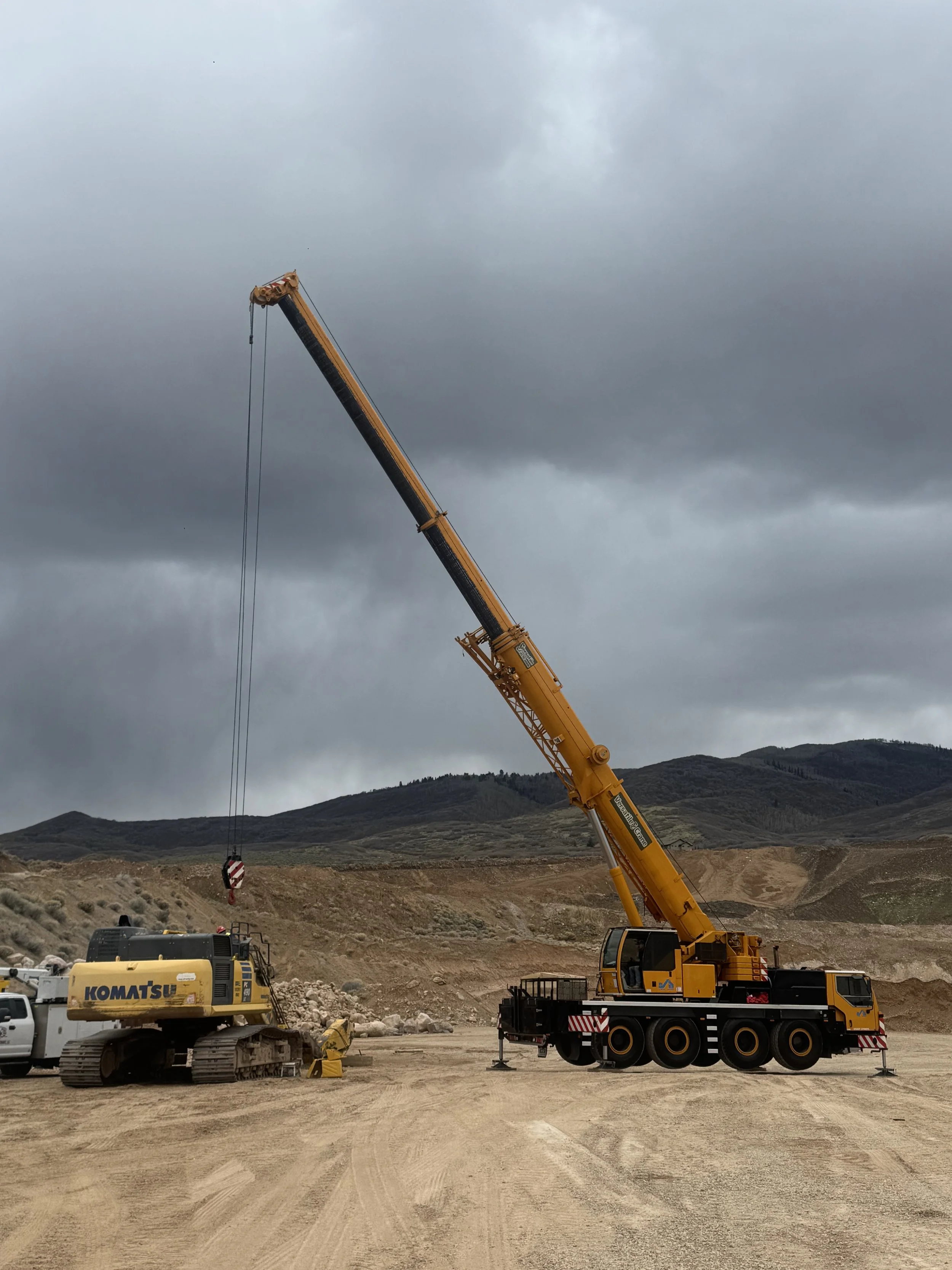 Construction site with a yellow crane lifting a load and a Komatsu excavator in the foreground, set against a backdrop of mountains and cloudy sky.