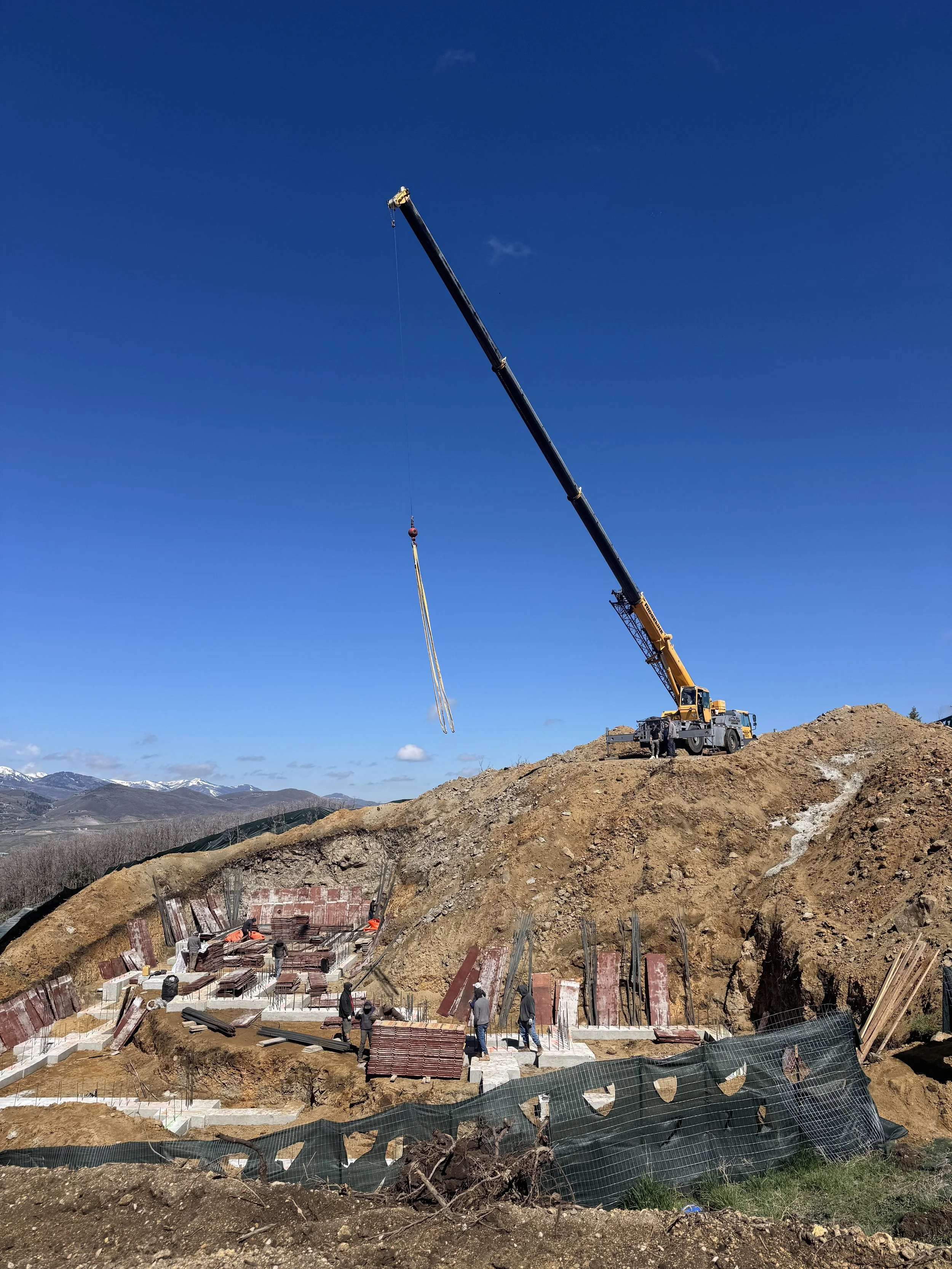 Construction site with a crane lifting materials, workers on site, and a hilly landscape in the background under a blue sky.