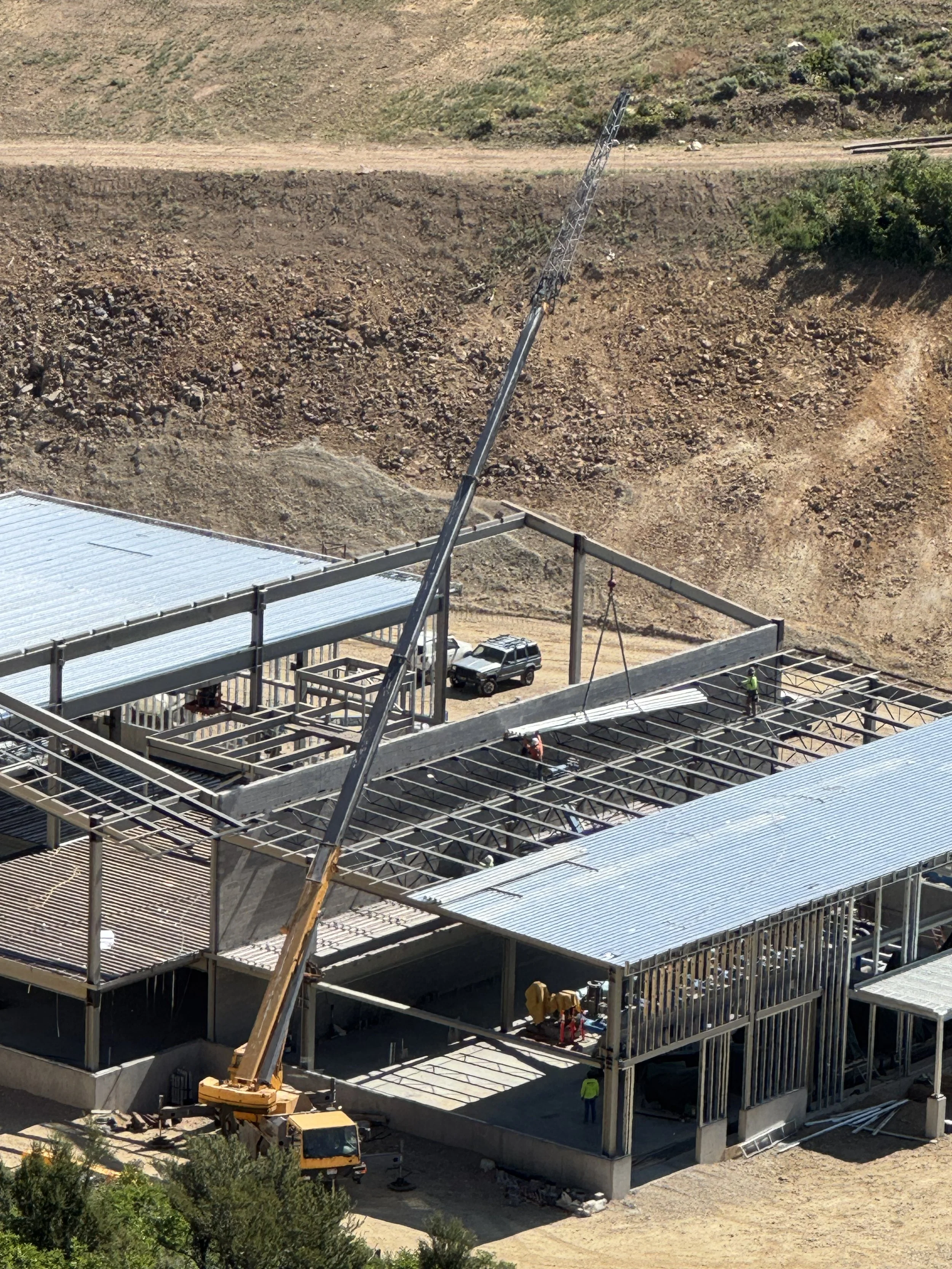 Construction site of a building with scaffolding, a crane, workers, and a partially completed roof amid a dirt landscape.