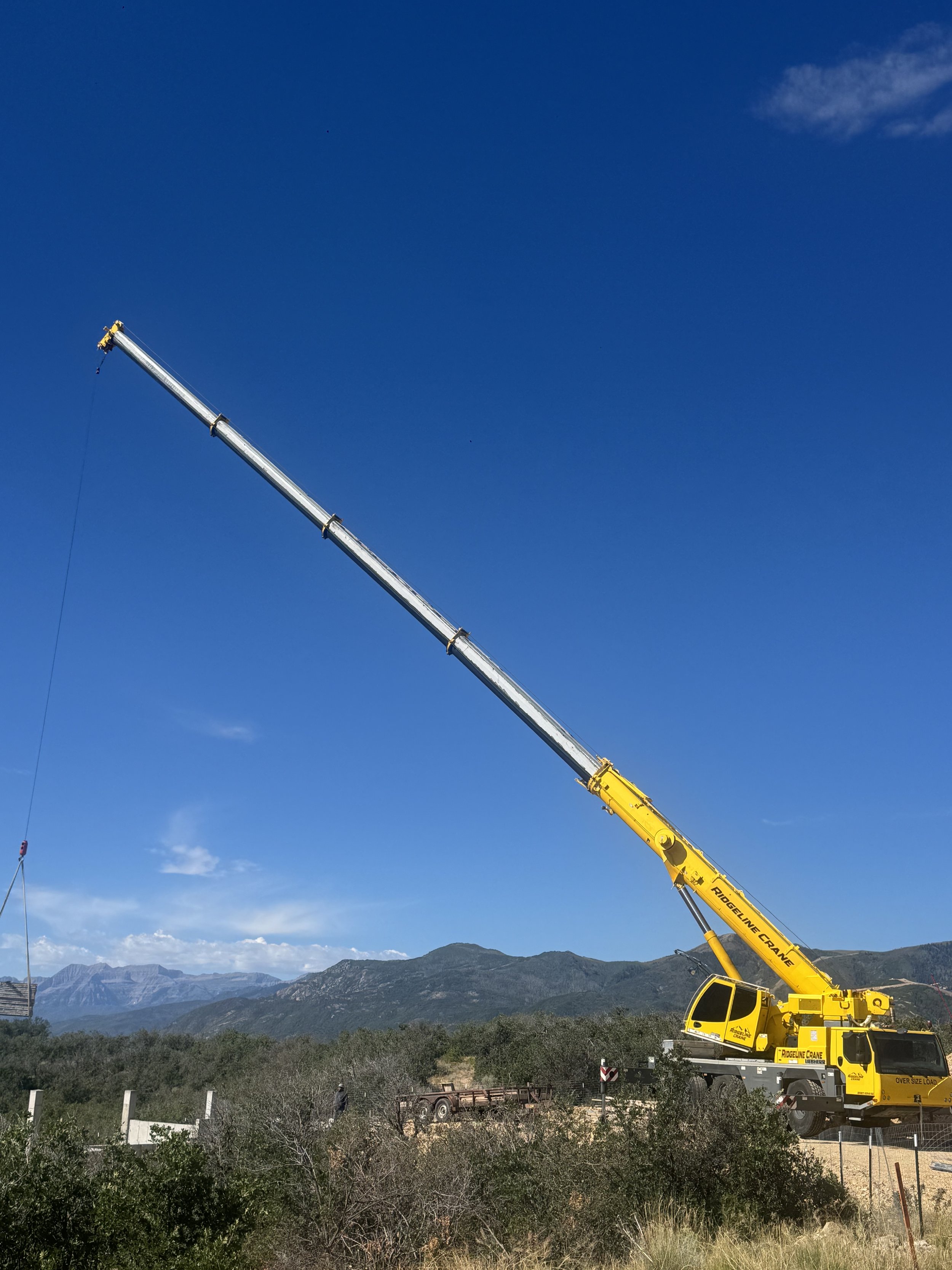 Large yellow crane with extended arm operating in a mountainous landscape, with green bushes and distant mountains under a blue sky with a few clouds.