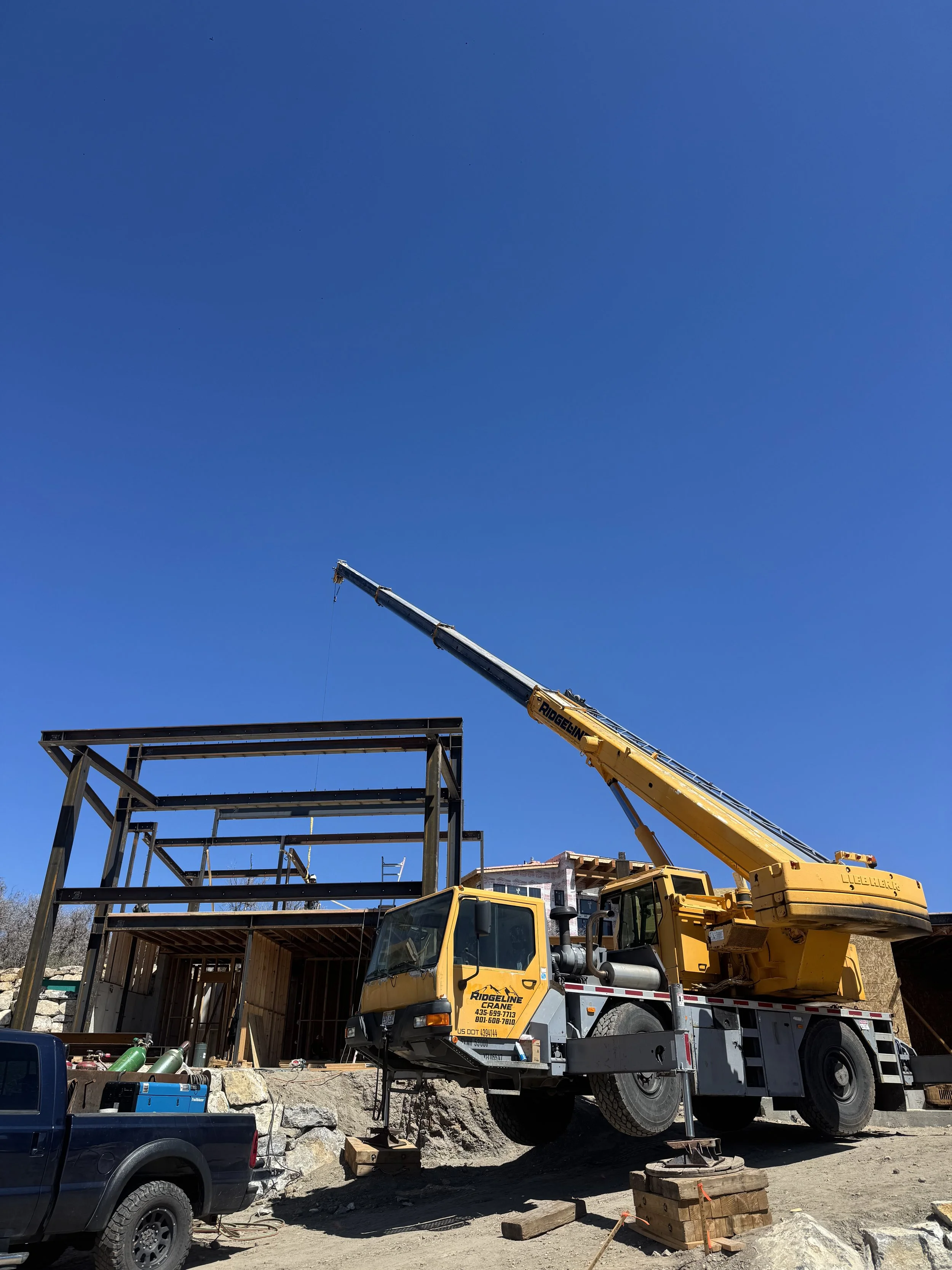Construction site with a yellow crane lifting a steel framework to build a structure, and vehicles parked nearby under a clear blue sky.