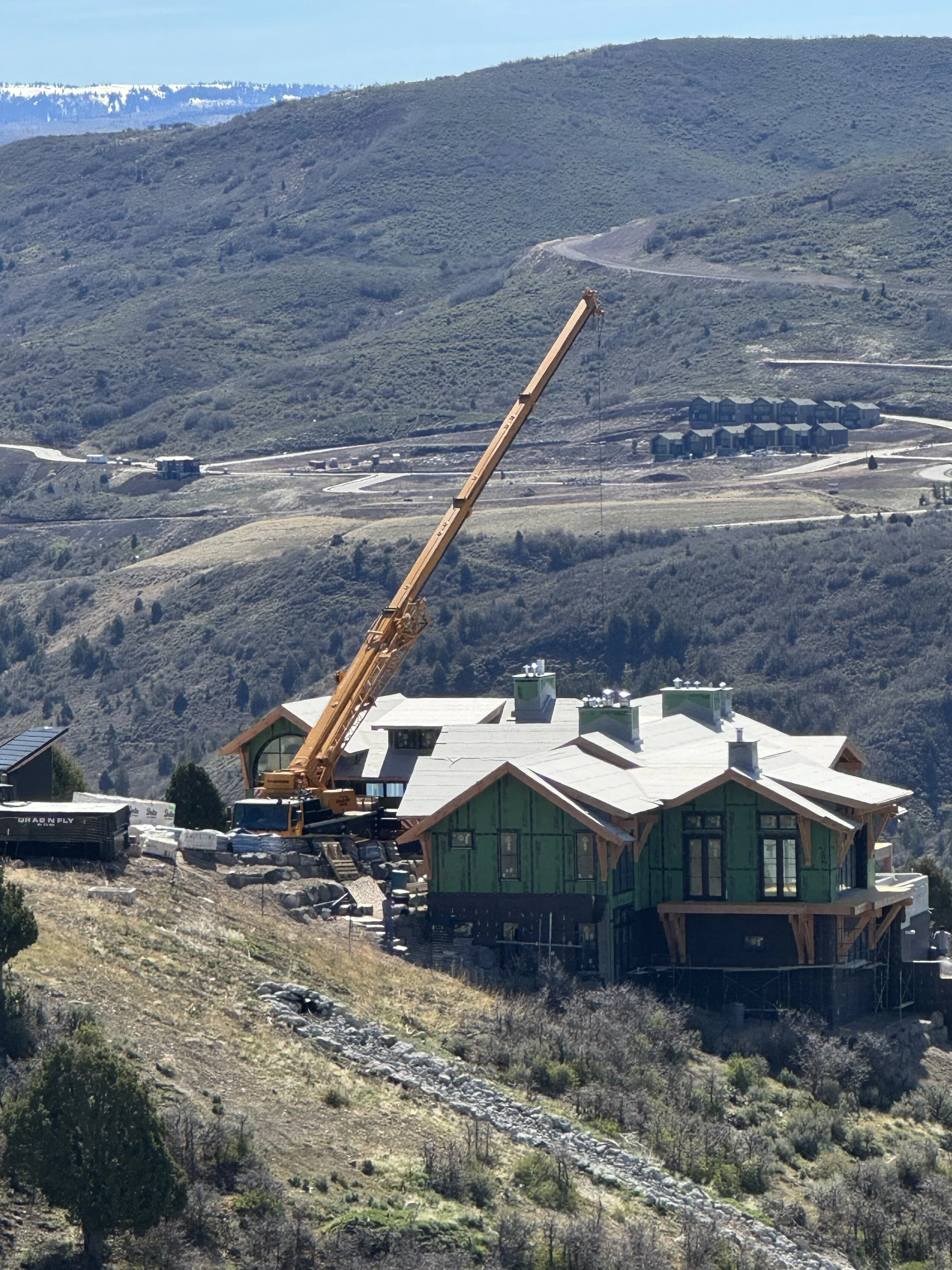 Construction site on a hillside with a large crane lifting materials, Fox Farmhouse-style house under construction, mountainous landscape in the background.