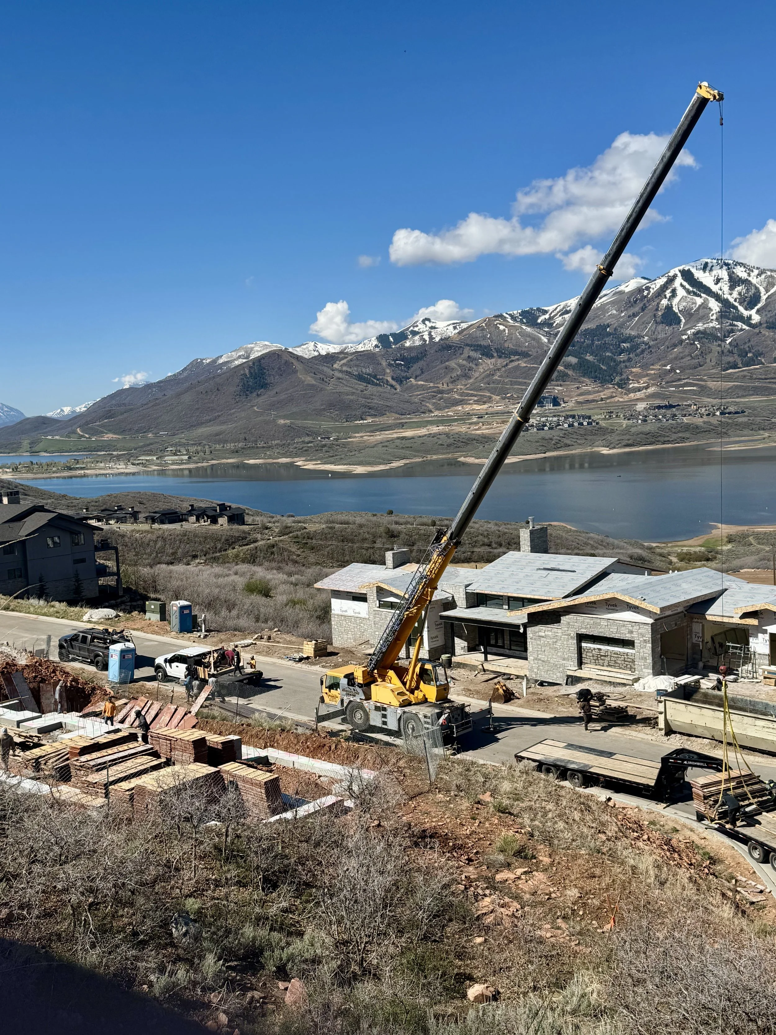 Construction site with a yellow crane lifting materials, houses under construction, a lake, and snow-capped mountains under a blue sky with clouds.