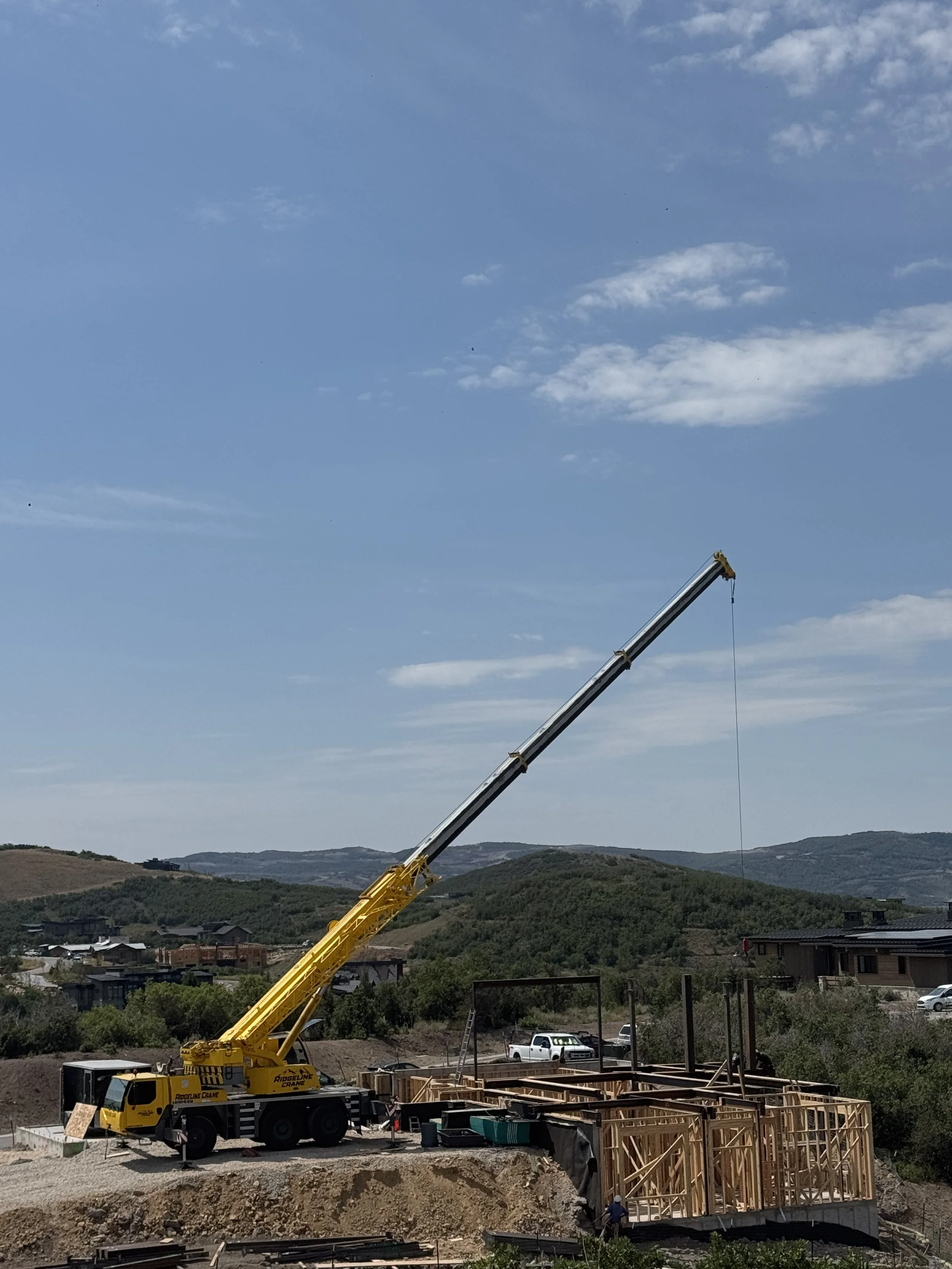 Construction site with a yellow crane lifting materials, wooden framing for a building, residential houses, green hills in the background, and a partly cloudy sky.
