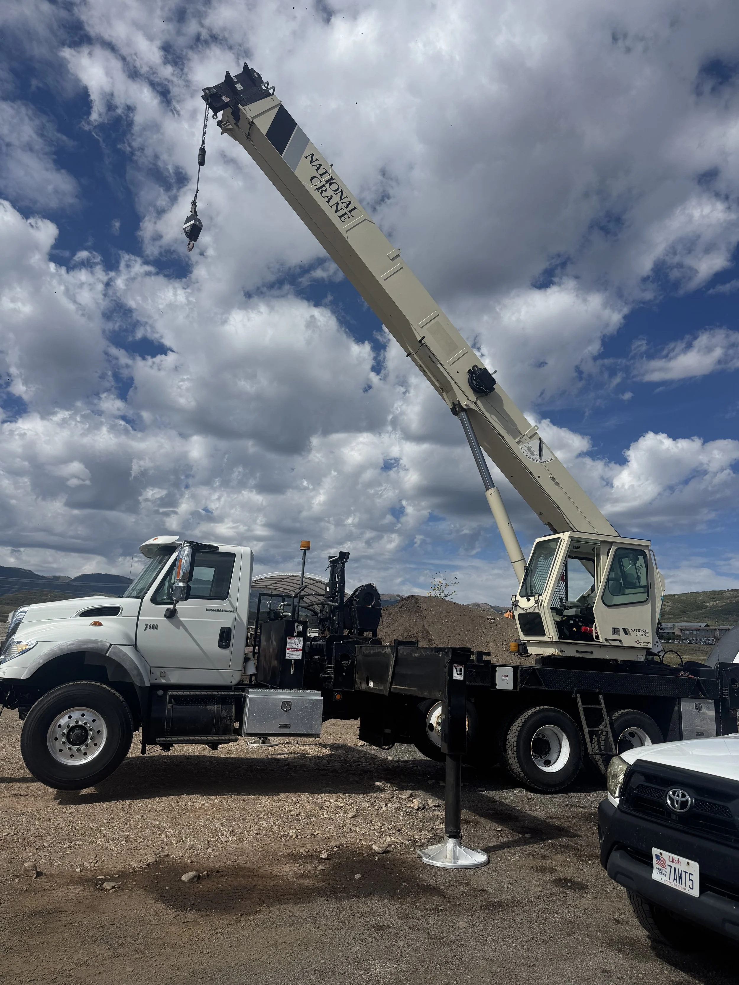 A large white crane truck with the label 'National Crane' on the arm, lifting a hook, parked on dirt ground under a partly cloudy sky. Other vehicles are visible nearby.