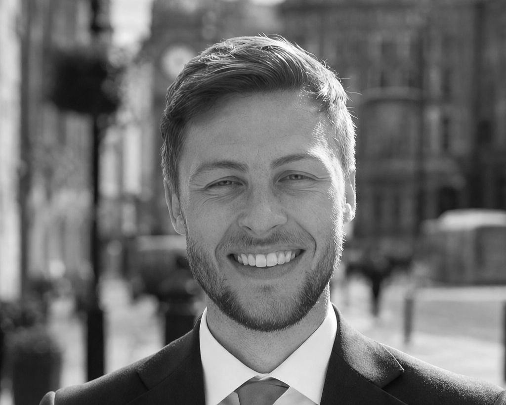 A man in a white shirt and dark tie smiling while sitting at a table with a glass of water, against a brick wall background.