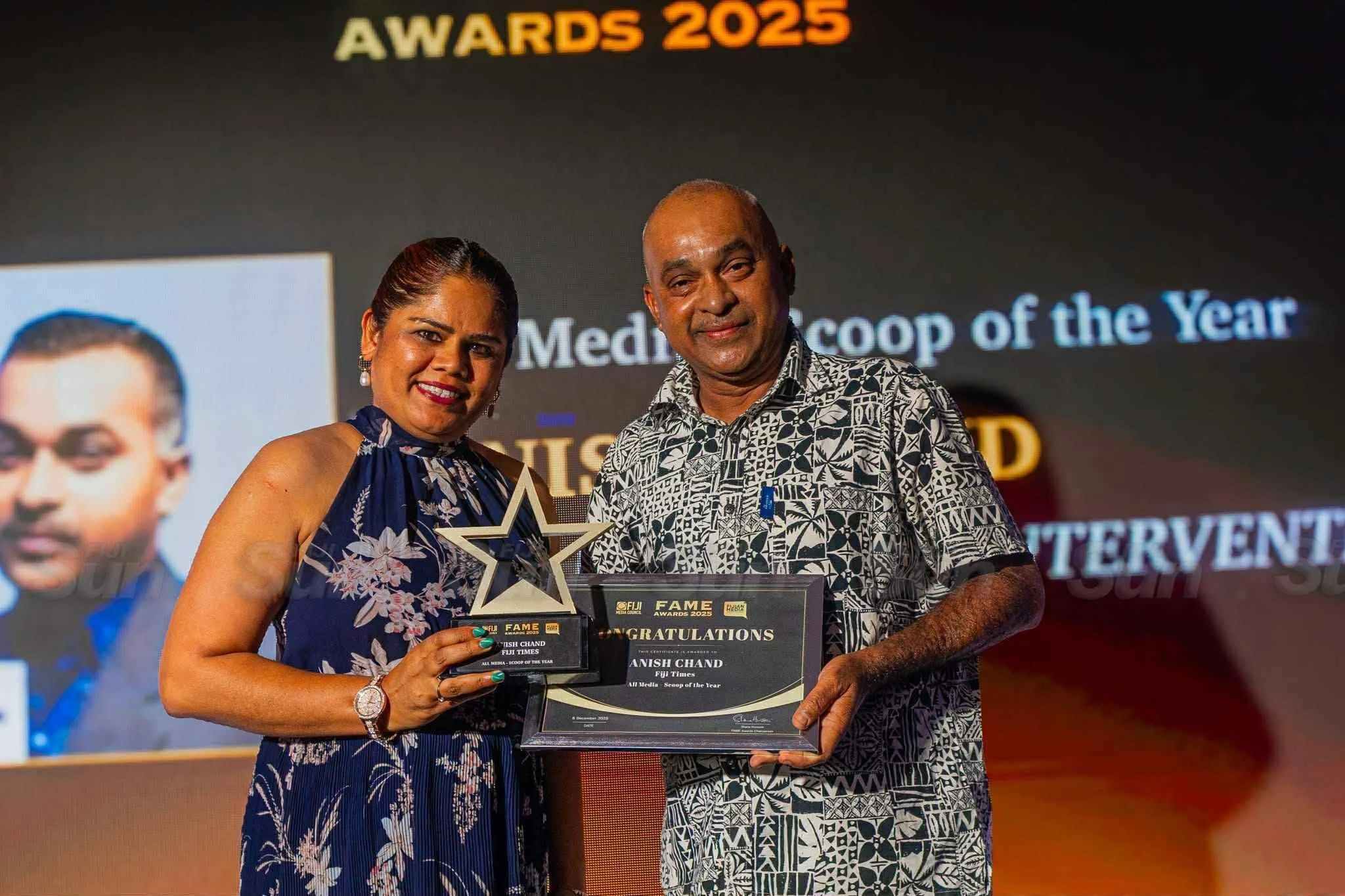 A woman and a man are holding awards and a certificate at an event. The woman is wearing a blue floral dress and the man is wearing a black and white patterned shirt. They are smiling and standing in front of a screen that says "Awards 2025" and "Media Scoop of the Year."