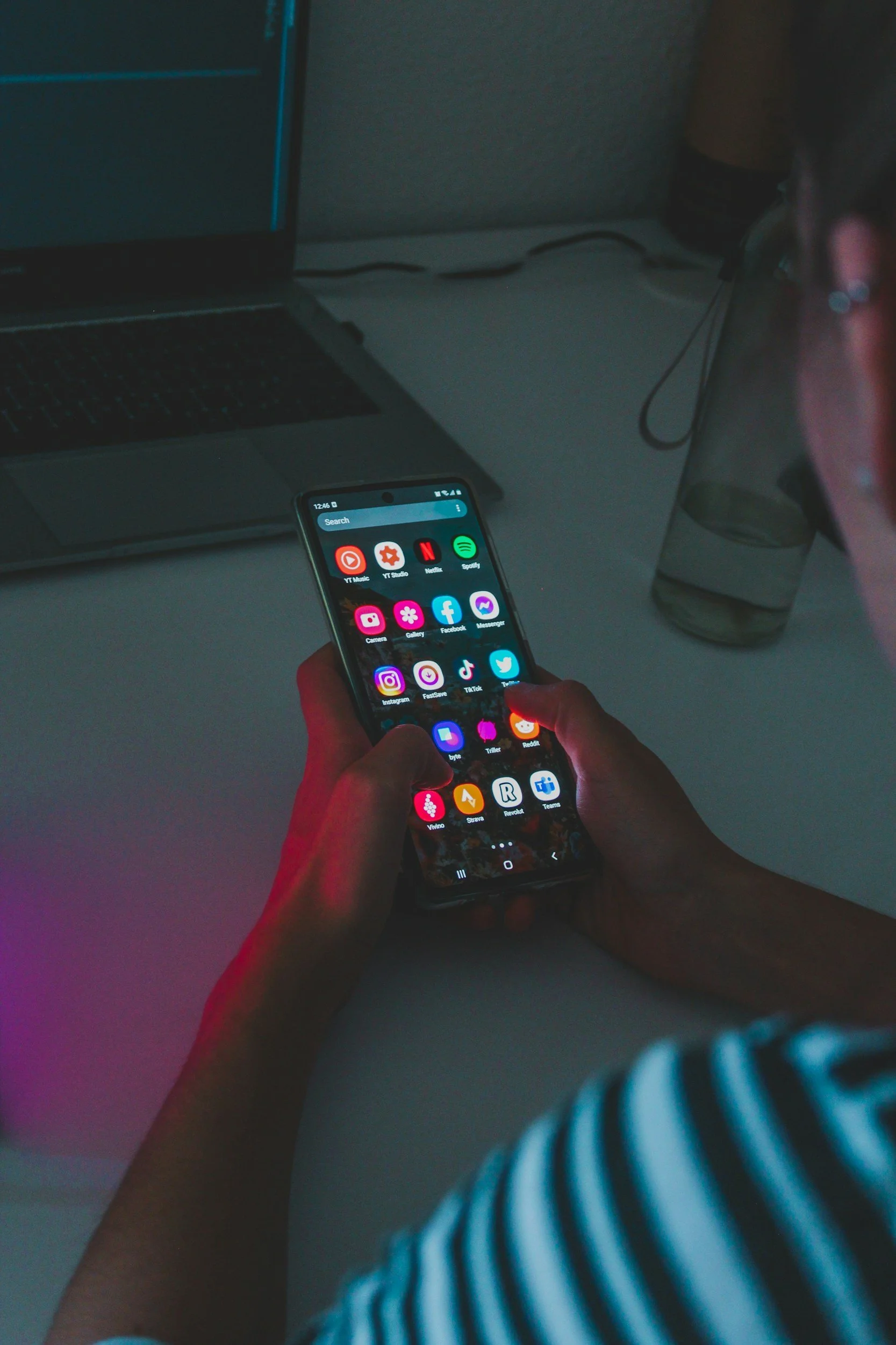 Person holding a smartphone with social media and app icons on the screen, sitting at a desk with a laptop and a glass of water nearby.