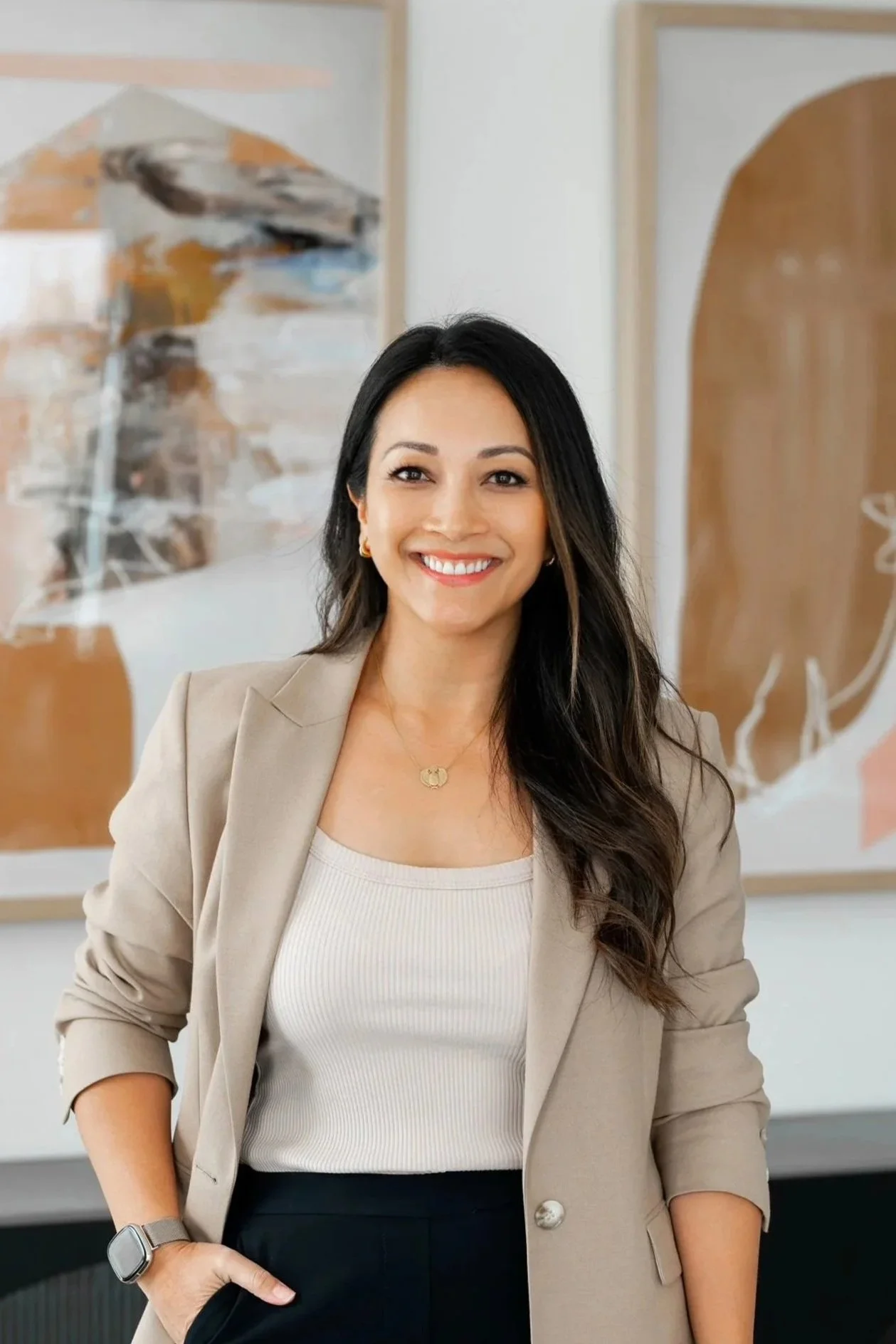 A woman with long dark hair, wearing a beige blazer over a white top, standing indoors with abstract art paintings in the background, smiling at the camera.