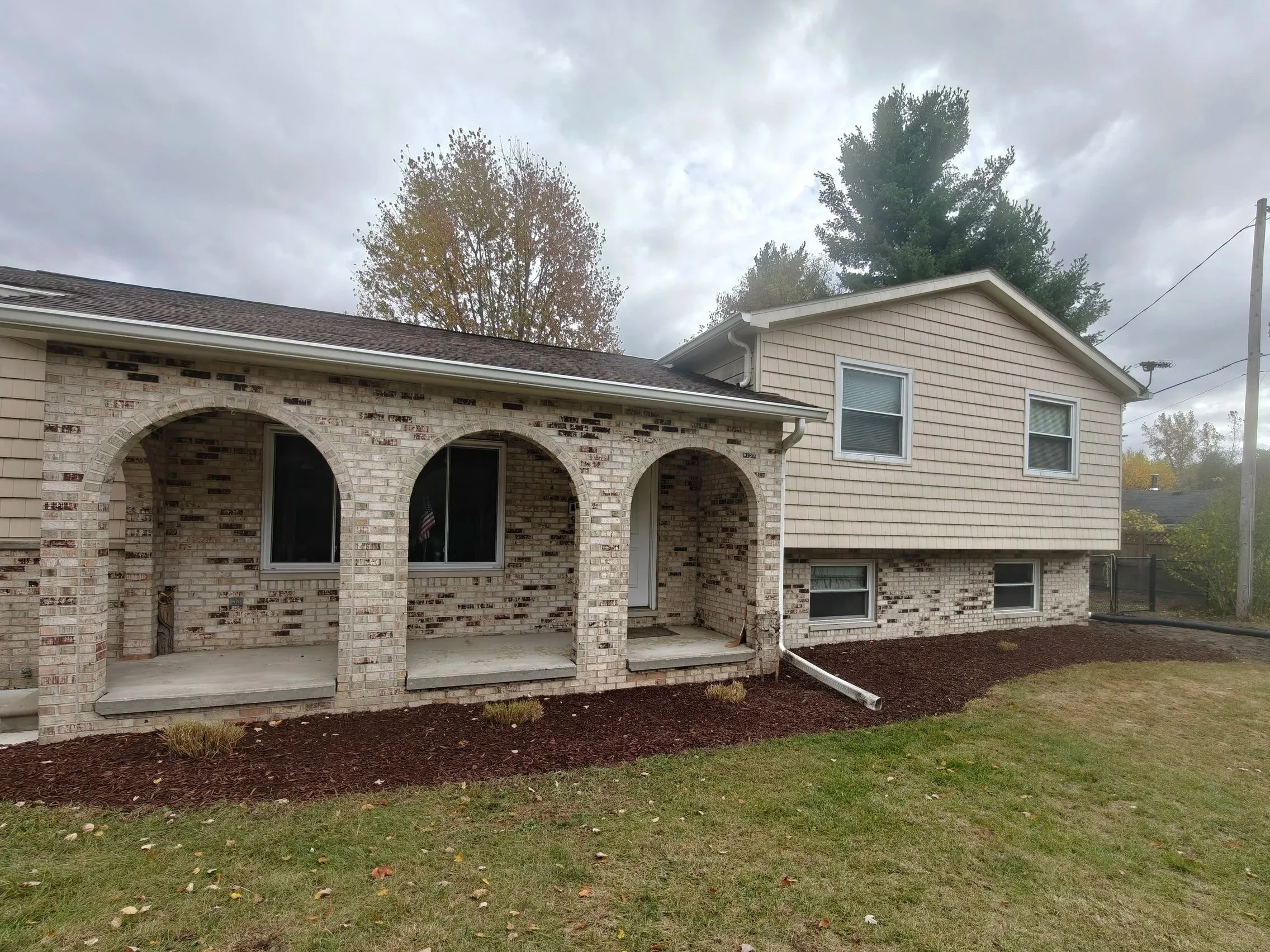 The image shows the back of a two-story house with a brick and beige siding exterior, three arched doorways, a small covered porch, and a freshly mulched yard area, under a cloudy sky.