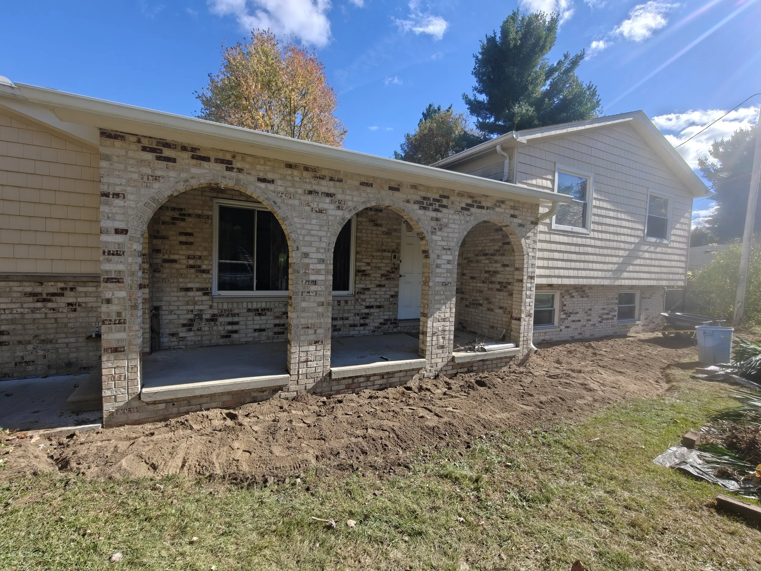 A house with a brick porch and three arches under construction, with bare dirt in the yard and a wheelbarrow and trash cans nearby.