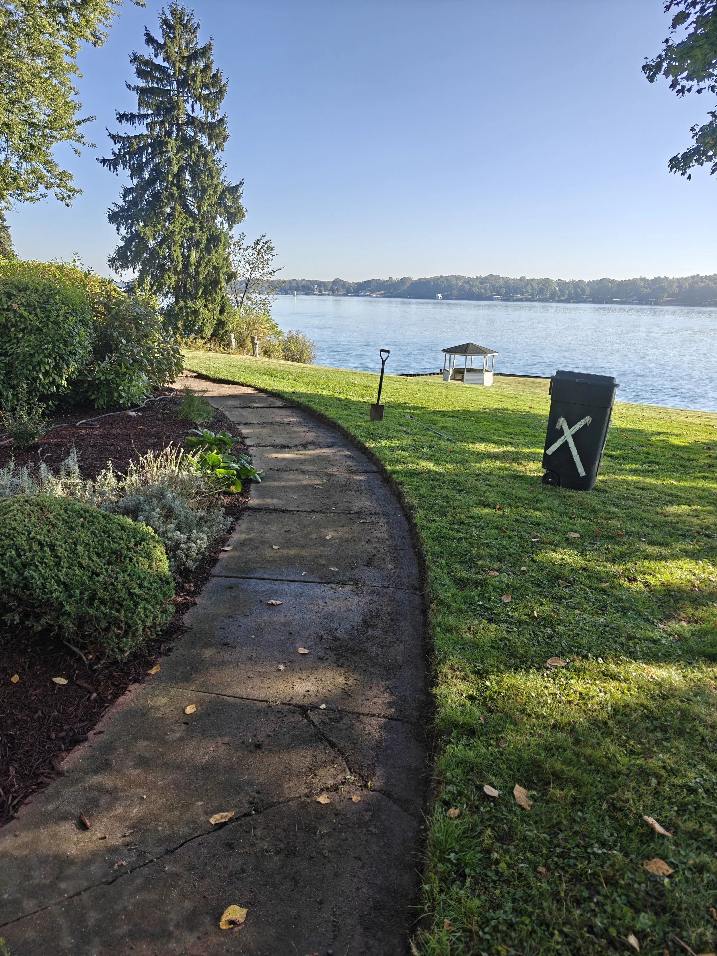 A lakeside park path with a garden on the left, a grassy area on the right, a garbage bin marked with a large X, a small gazebo over the water, and trees in the background on a clear, sunny day.