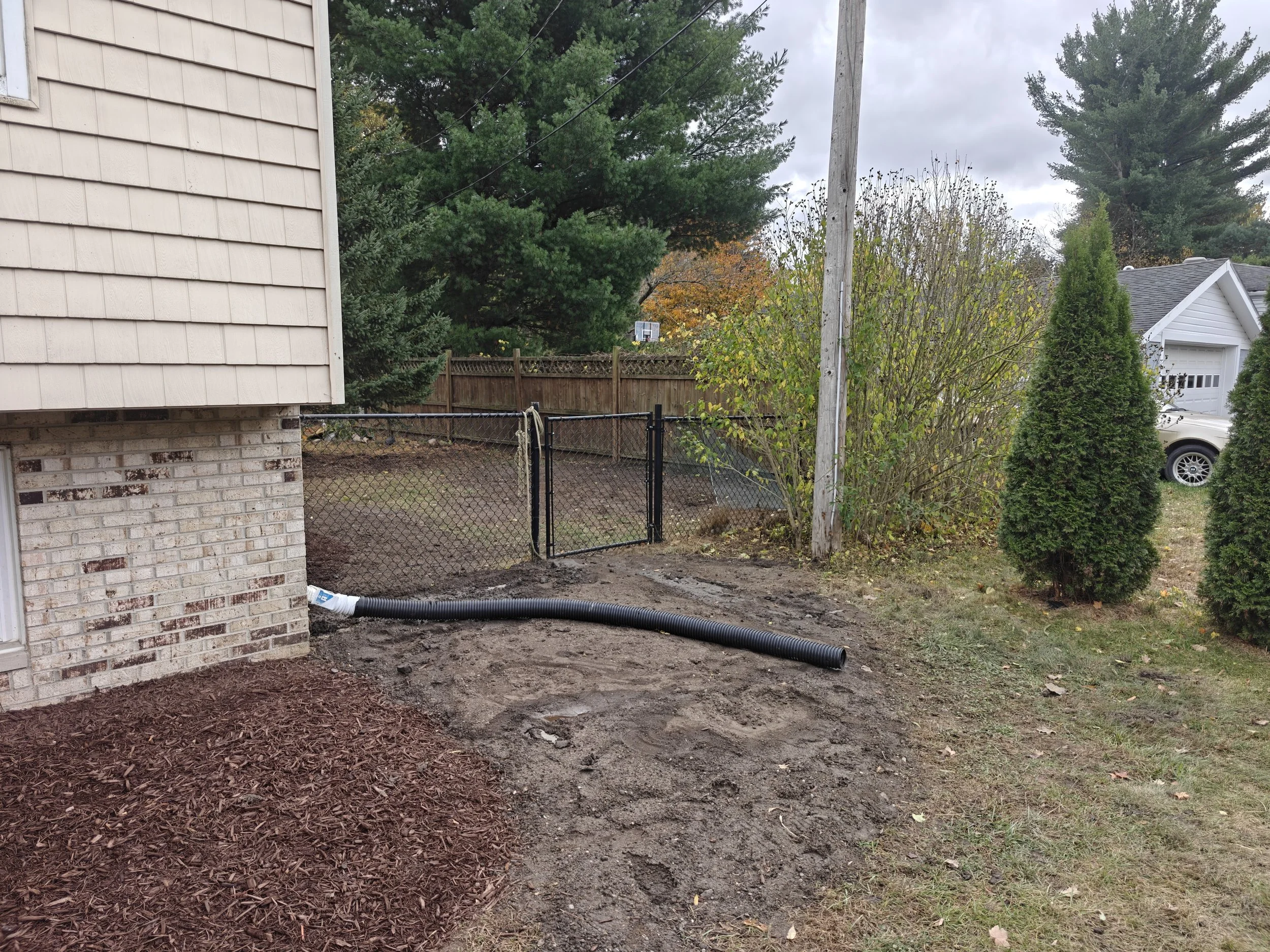 View of a backyard with a house on the left, a chain-link fence with a gate, a utility pole, and various trees and shrubs. The ground appears to be disturbed soil with a black drainage pipe laying on the ground.