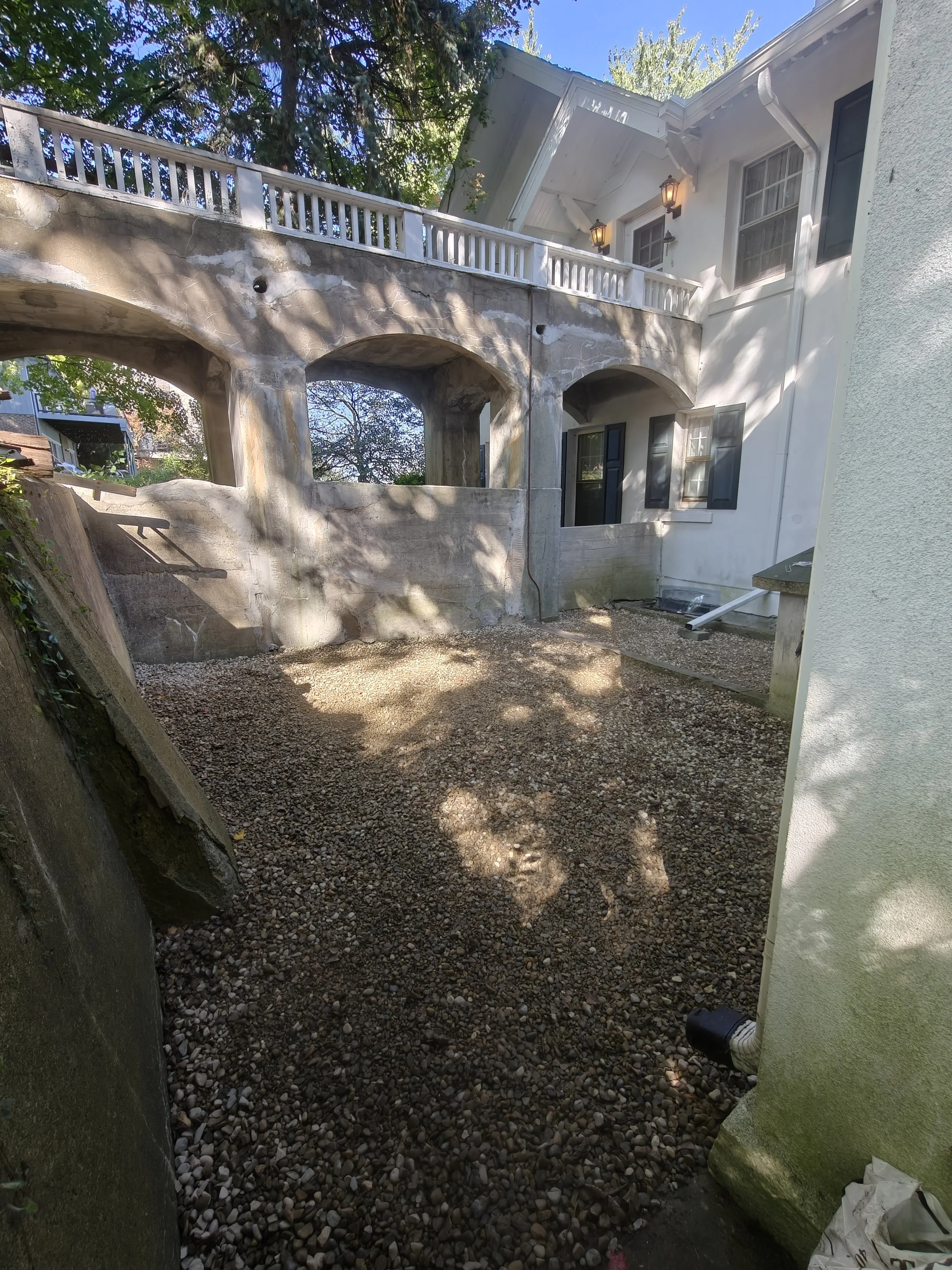 Backyard with gravel ground, stone walls, and a white house with dark shutters, outdoor lighting, and a patio area.