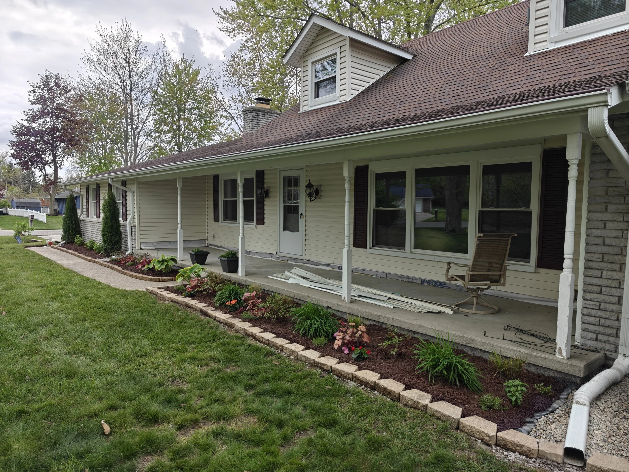 Side view of a house with a new porch renovation. The house has beige siding, dark shutters, and a brown roof. The porch has some construction materials and a chair, with an unfinished look. A flower bed with blooming plants runs along the porch edge