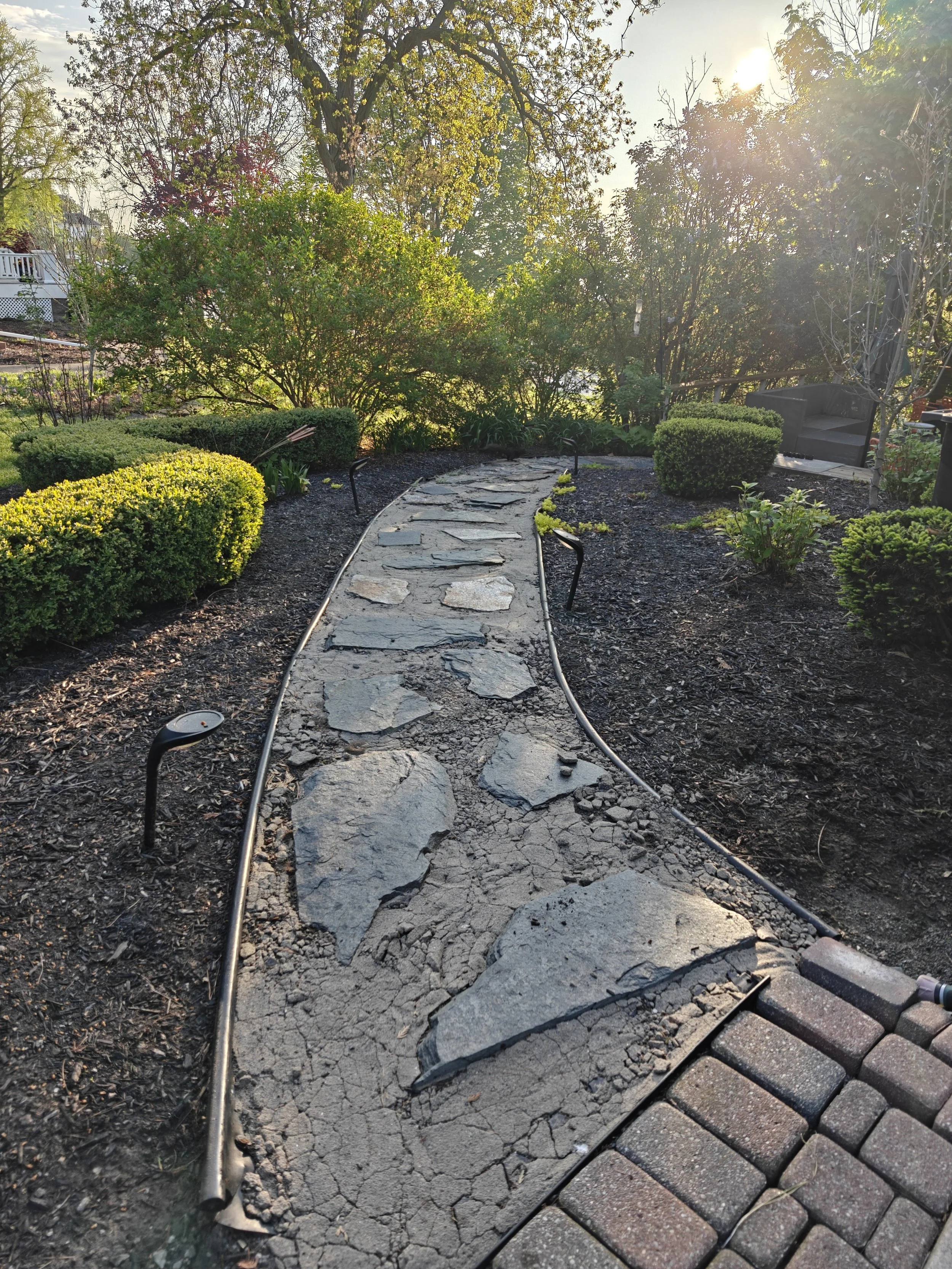 A garden pathway under repair with scattered broken flagstone stones, surrounding green bushes, and a sunlight shining through trees in the background.
